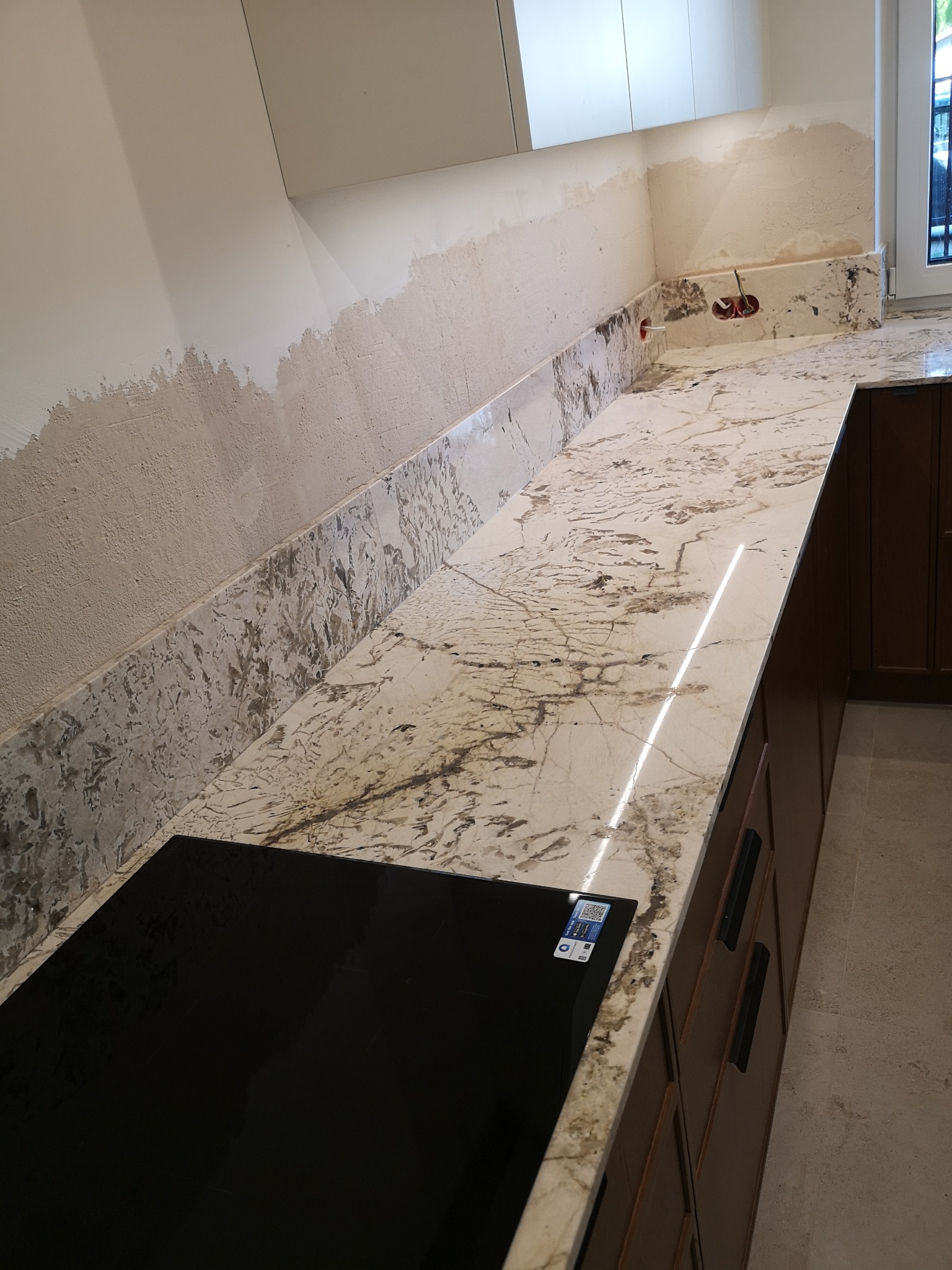 Montaż blatu kamiennego w kuchni. Widoczne niedokończone prace wykończeniowe na ścianie. Na blacie płyta indukcyjna. Uwagę przykuwa unikalny wzór kamienia.