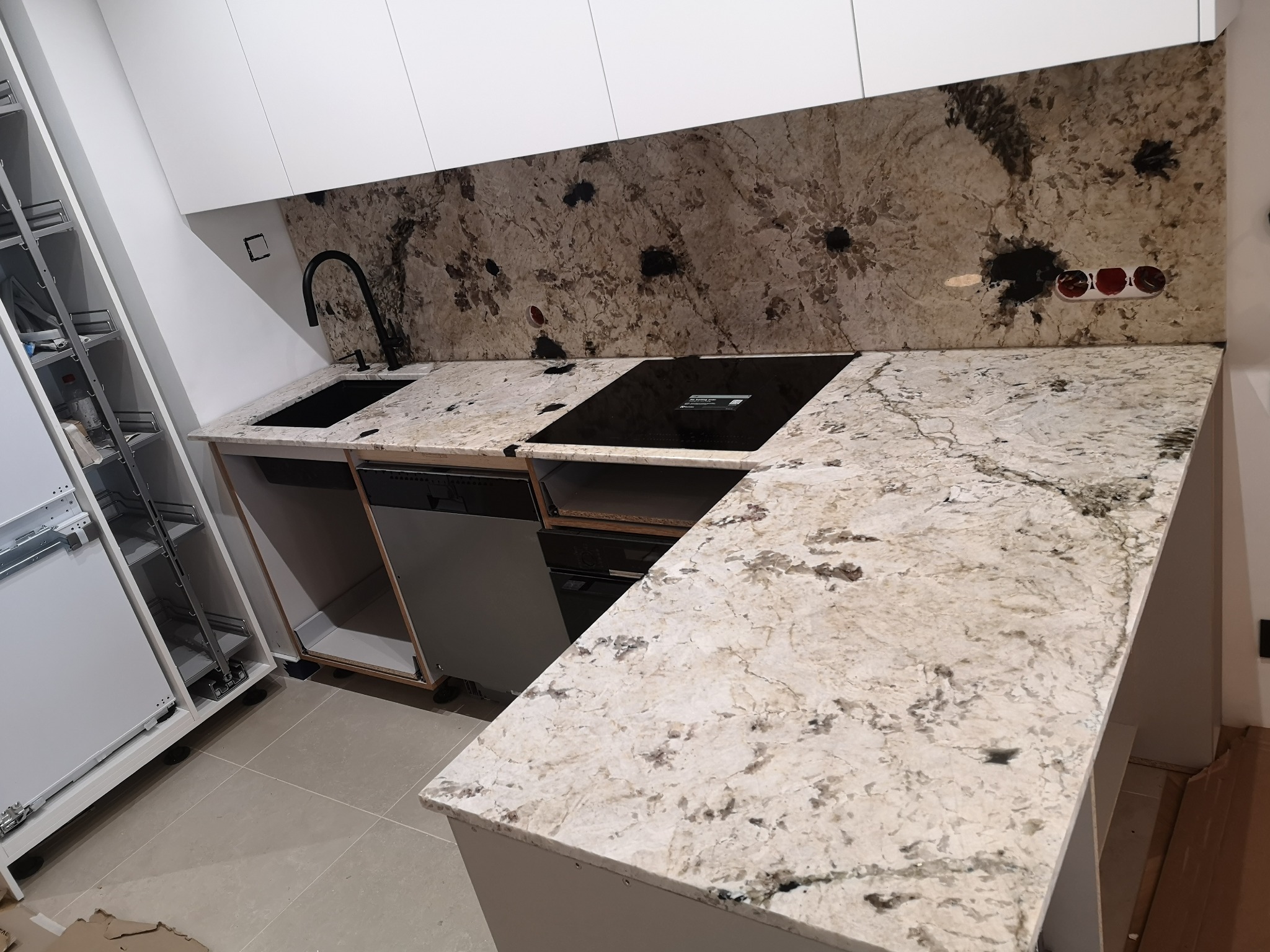 Jasny blat z kamienia w kuchni z czarną armaturą i płytą grzewczą. Widoczny fragment ściany z kamiennym panelem i białe szafki kuchenne.