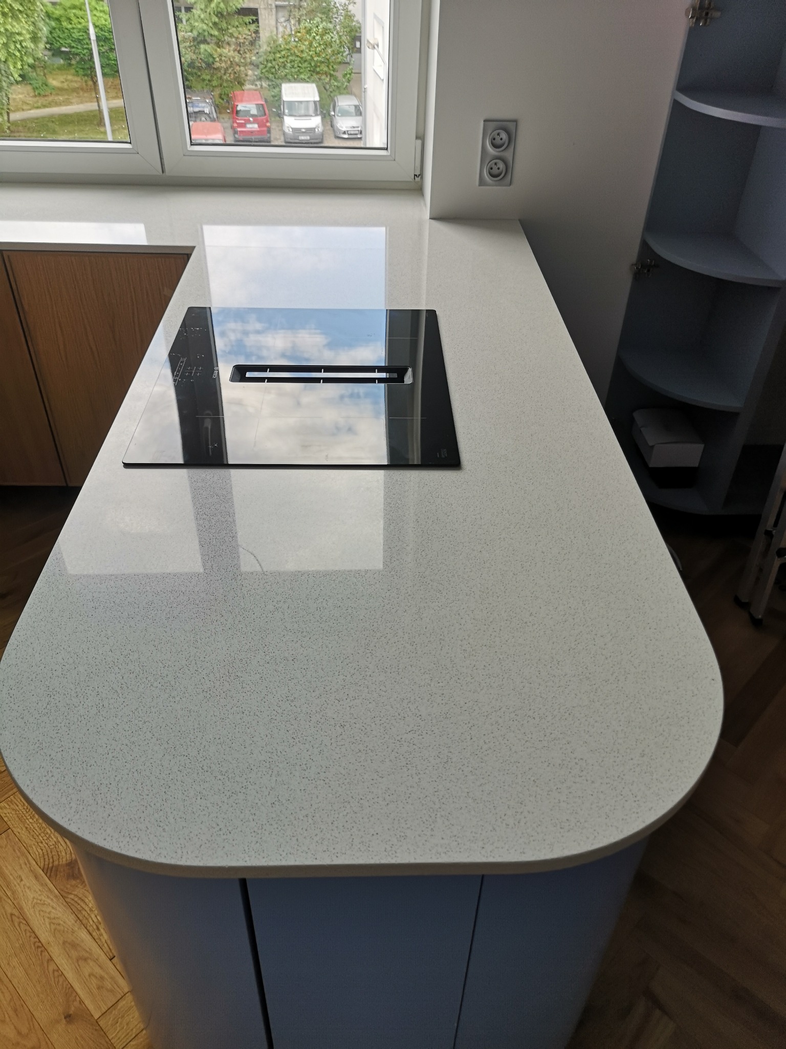 Jasny blat kuchenny z konglomeratu z wbudowaną płytą indukcyjną, odbijający niebo. Za oknem widok na zieleń i zaparkowane samochody.