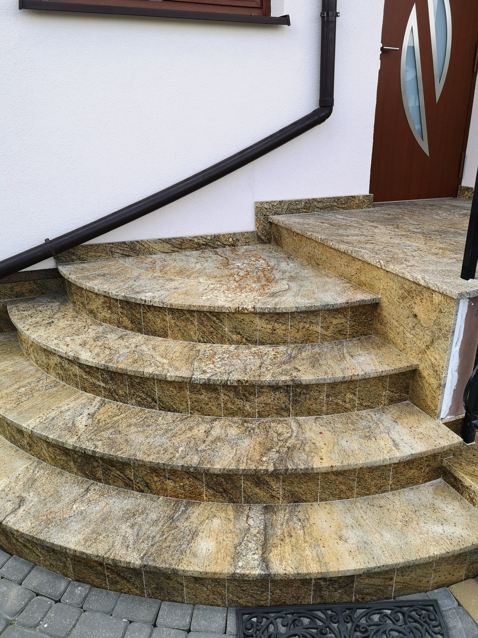 Zaokrąglone schody zewnętrzne z kamienia w odcieniach beżu i brązu, prowadzące do drzwi wejściowych. Widoczny fragment elewacji budynku i rynna.