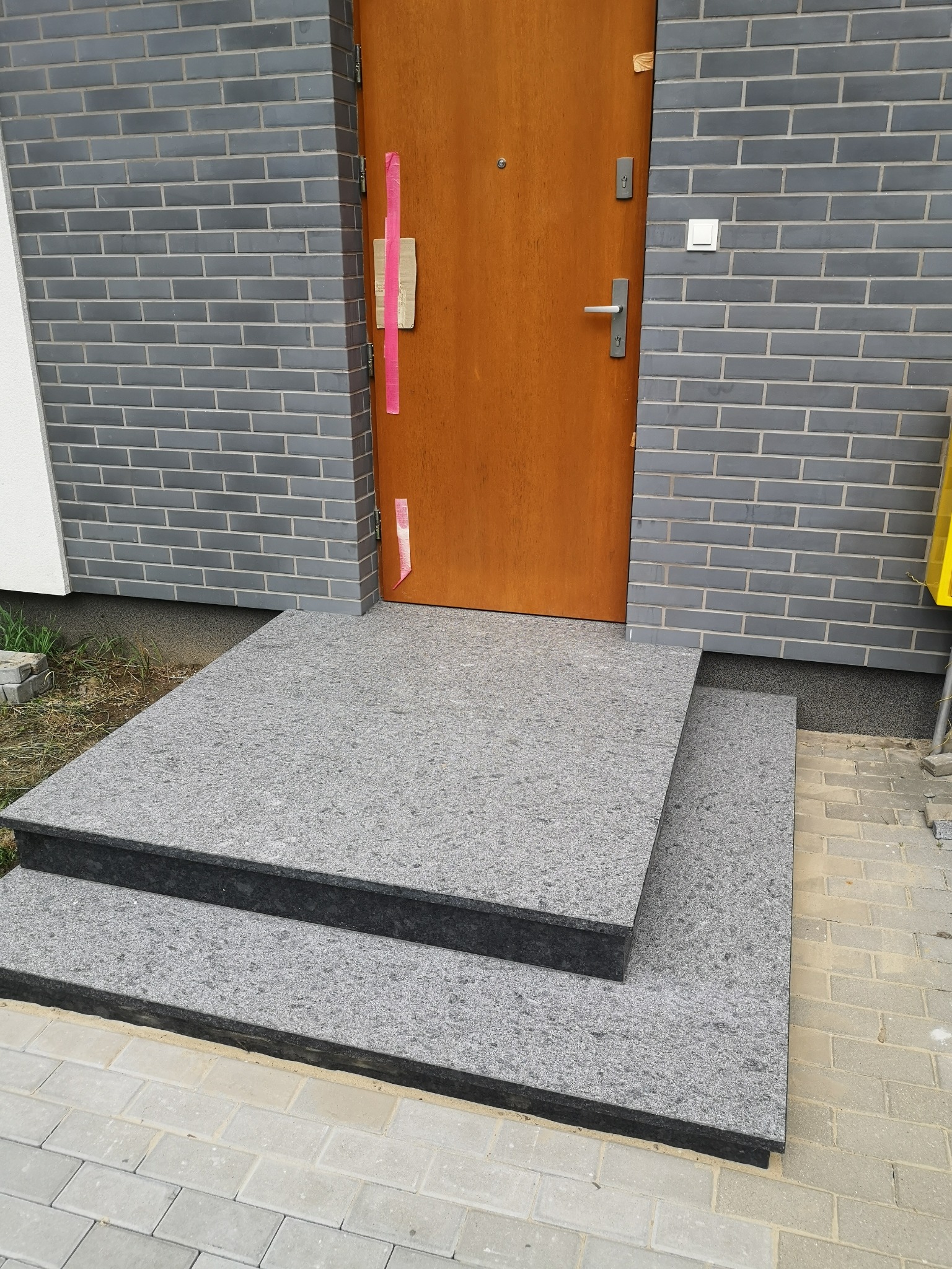 Dwu-stopniowe schody wejściowe z szarego kamienia, prowadzące do drewnianych drzwi w budynku z szarej cegły. Widoczny fragment chodnika z kostki.