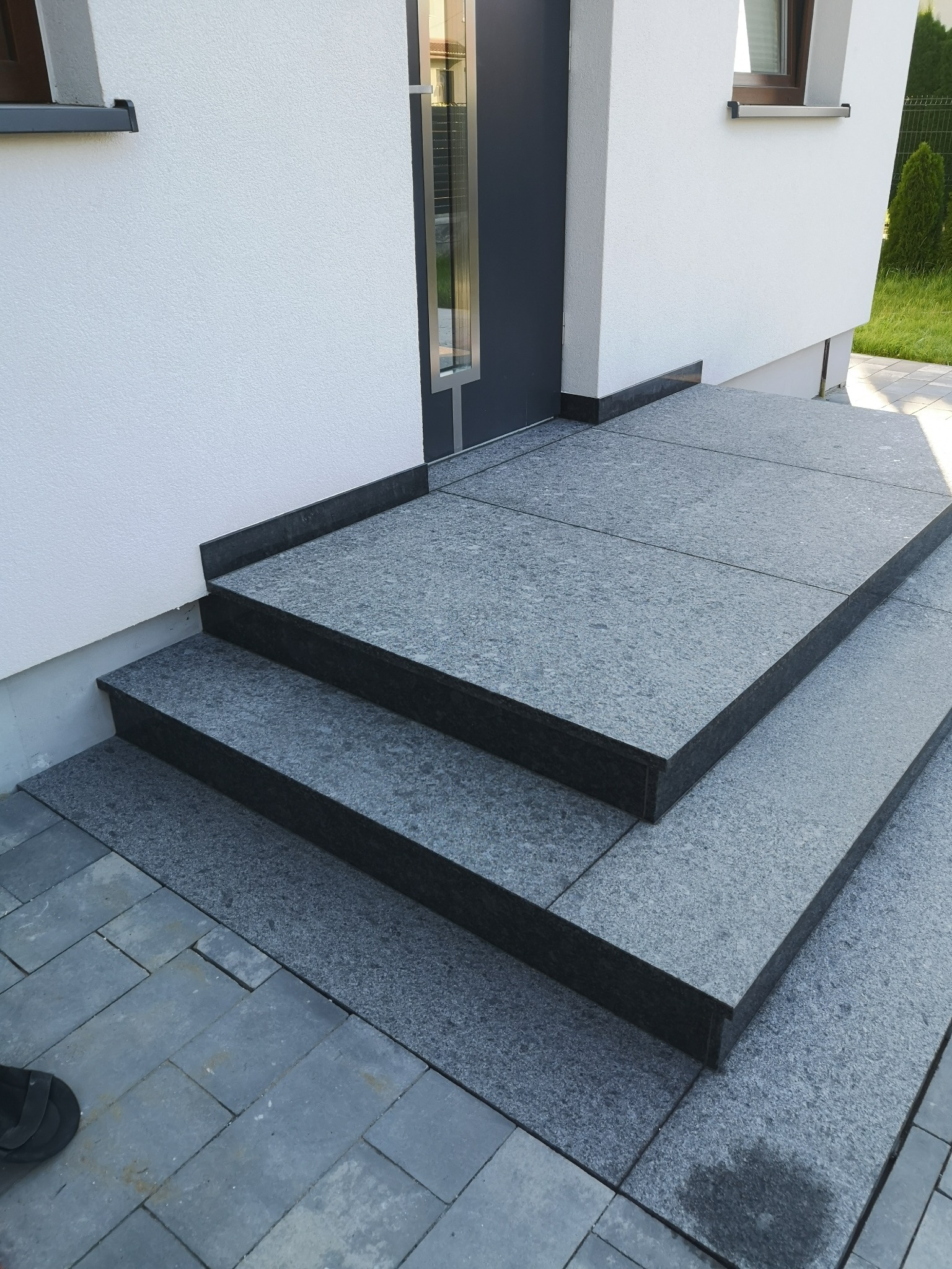 Zewnętrzne schody z szarego kamienia prowadzą do drzwi wejściowych, kontrastując z białą fasadą budynku. Widoczny fragment chodnika z kostki brukowej.