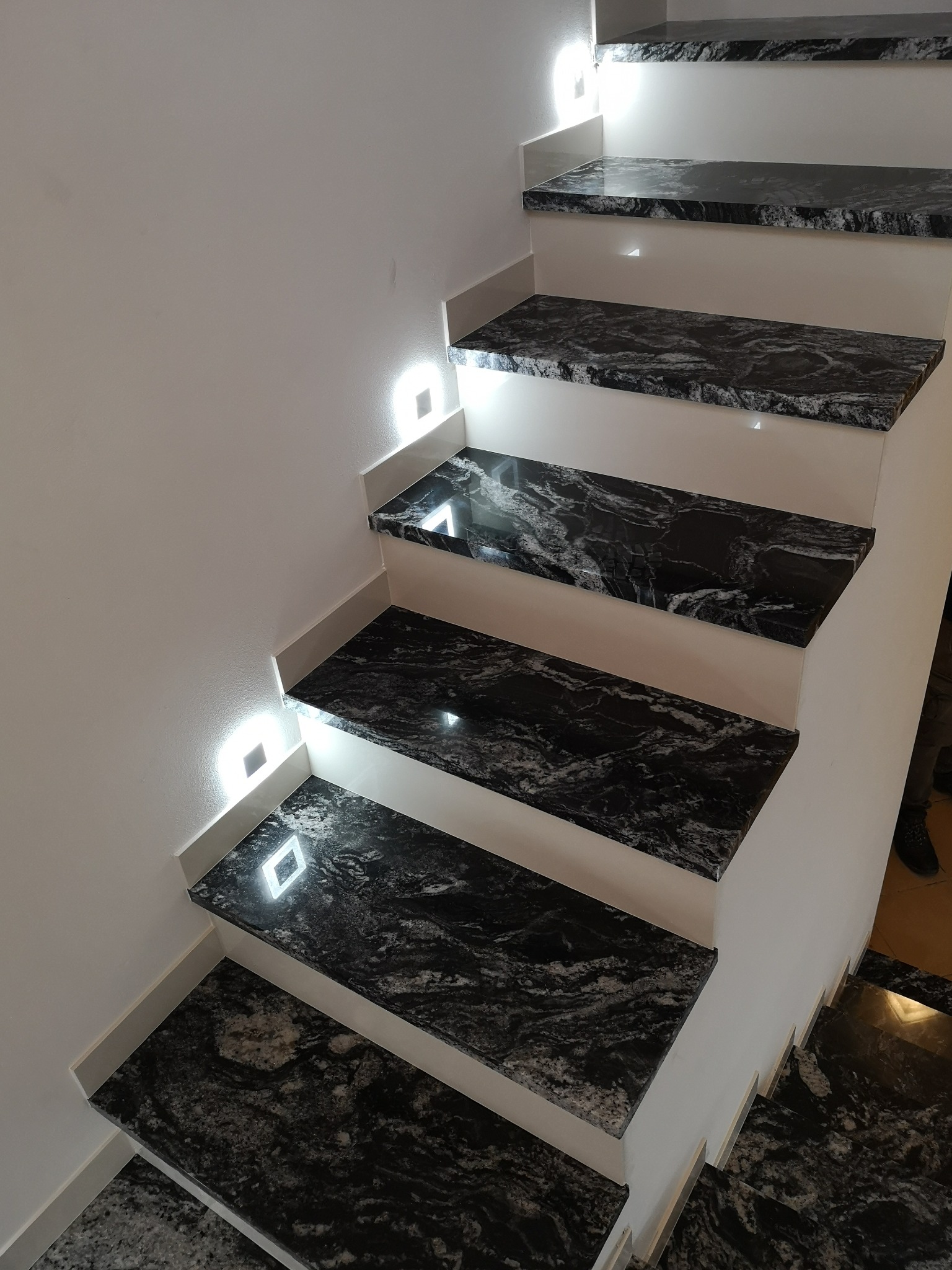 Schody kamienne z podświetleniem LED wzdłuż białej ściany, ukazujące unikalny wzór czarnego kamienia z białymi żyłkami i eleganckie wykończenie.