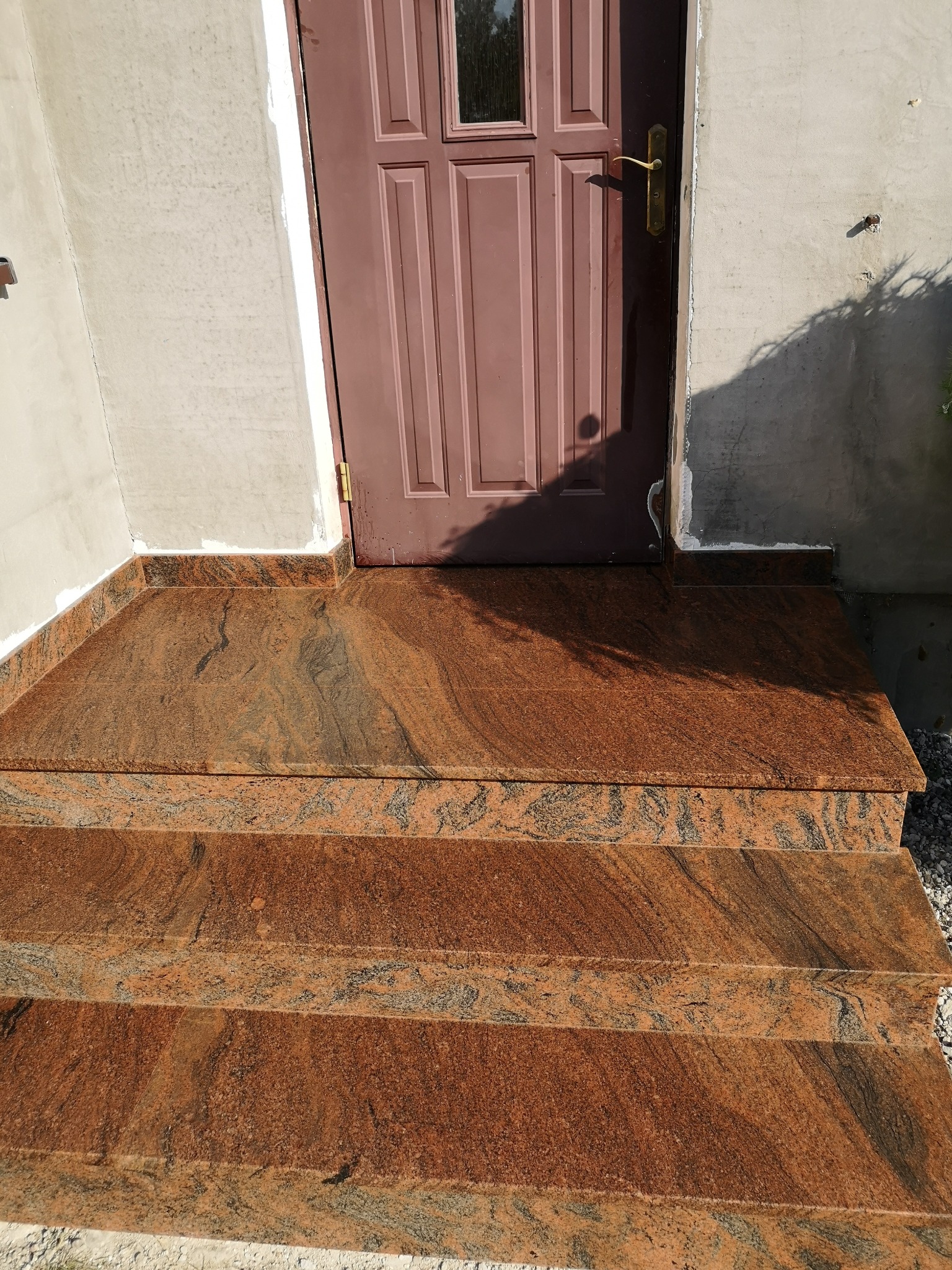 Zewnętrzne schody wejściowe z czerwonego granitu z widocznym wzorem. Trzy stopnie prowadzą do brązowych drzwi wejściowych. Ściana w tle szara, częściowo otynkowana.