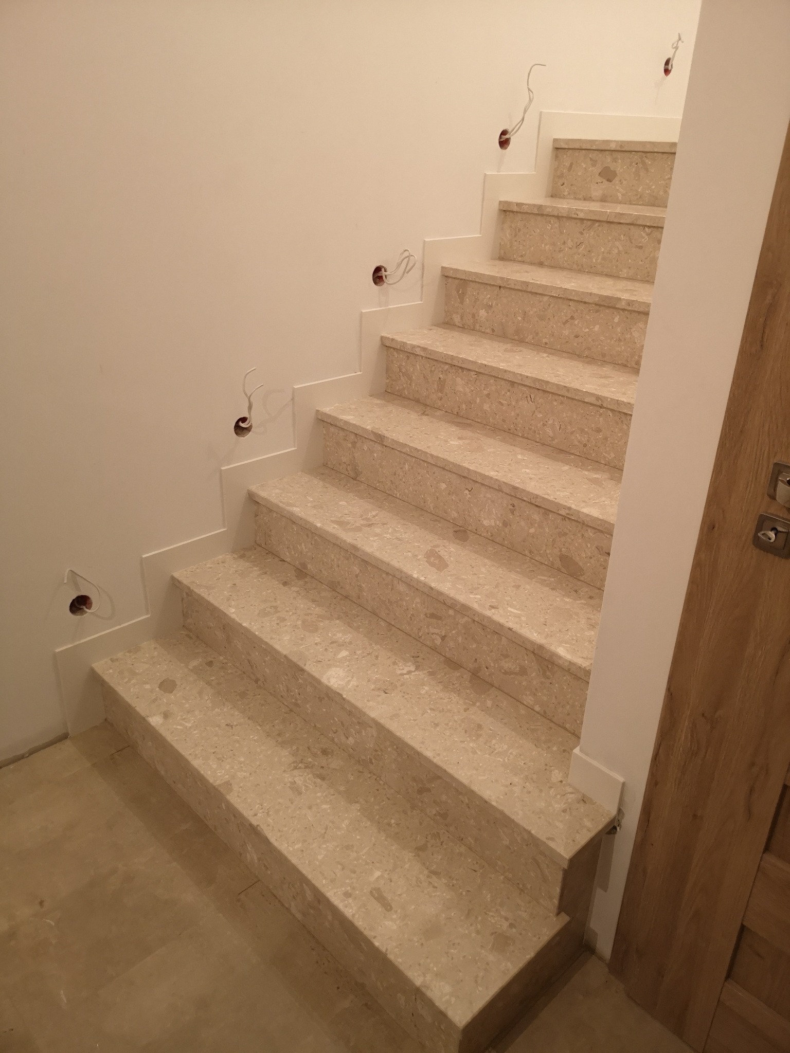 Jasne, wewnętrzne schody z terazzo, z widocznymi puszkami instalacyjnymi w ścianie, prowadzące do drewnianych drzwi.