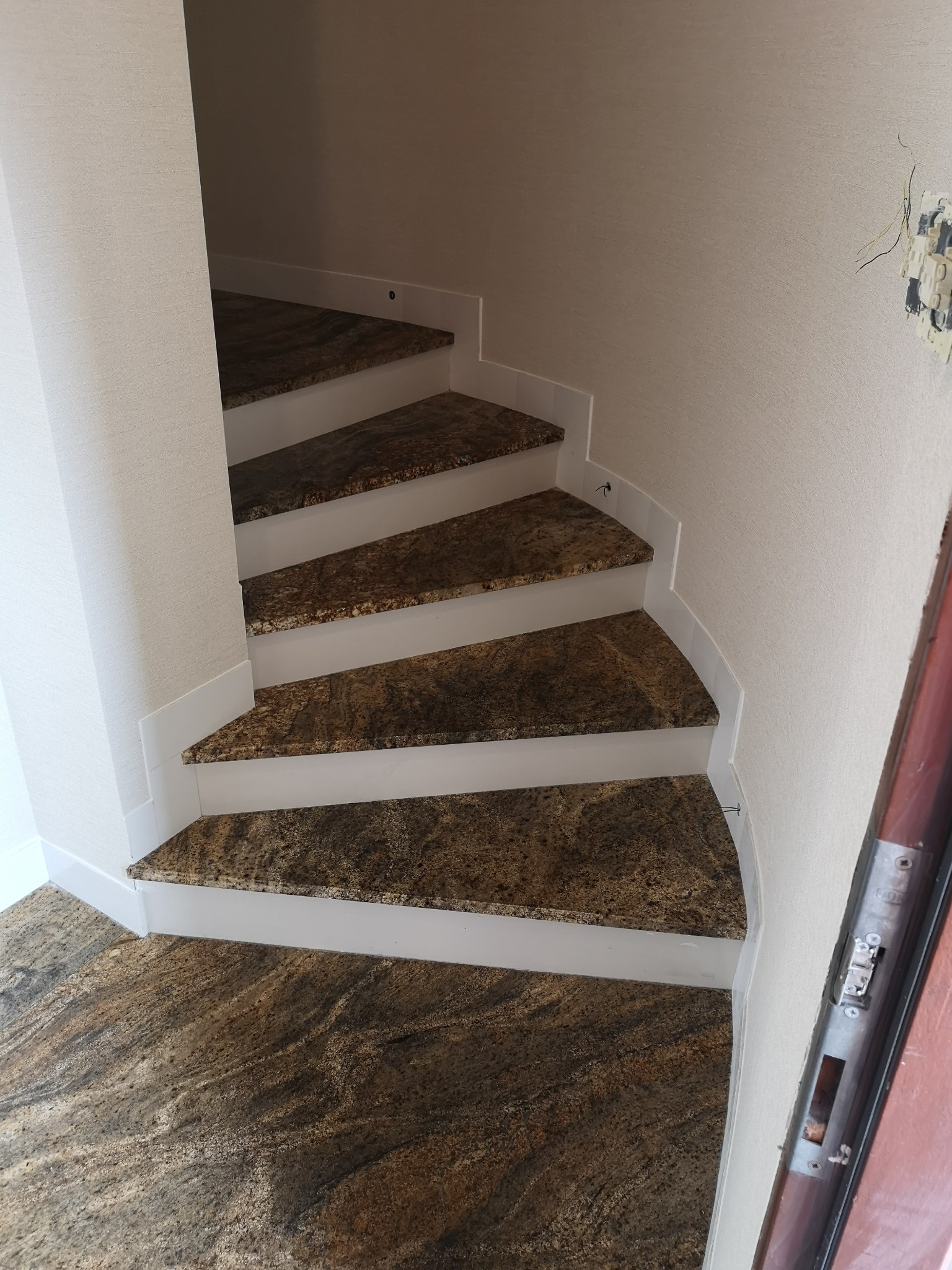 Wewnętrzne schody z brązowo-beżowego kamienia z białymi podstopnicami, widok z dołu na zakręt schodów, obok framuga drzwi i niedokończone gniazdko elektryczne.