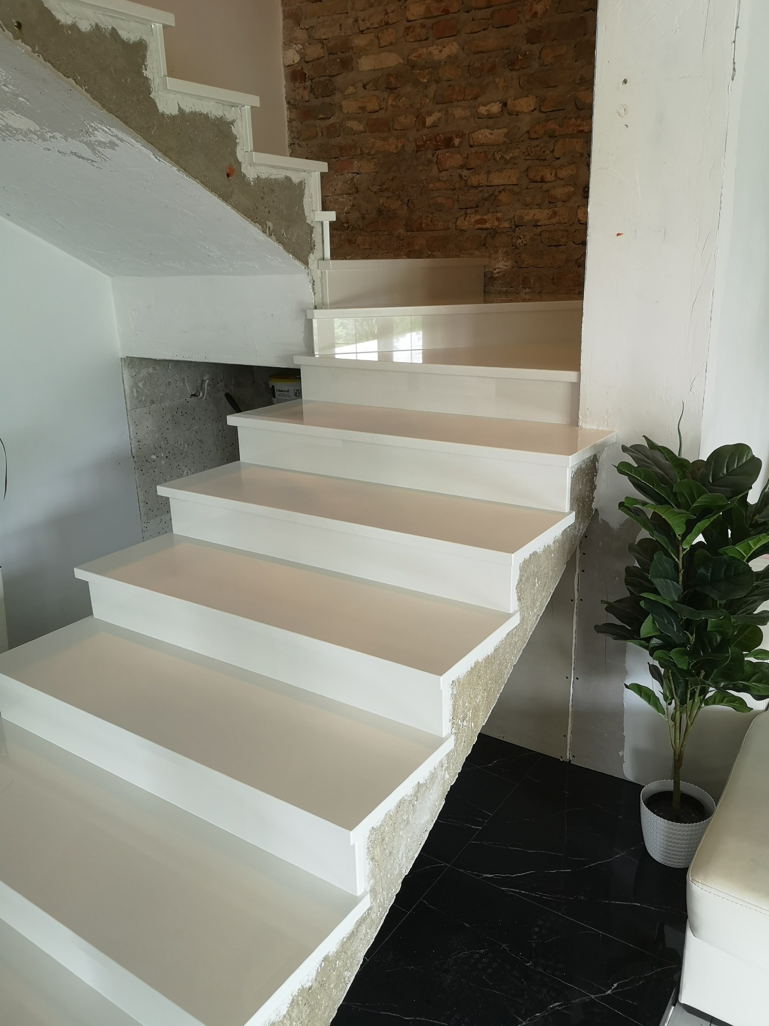 Białe schody kamienne w trakcie montażu, widoczne surowe wykończenie ściany i fragment ceglanej ściany, uzupełnione rośliną doniczkową na czarnej marmurowej podłodze.