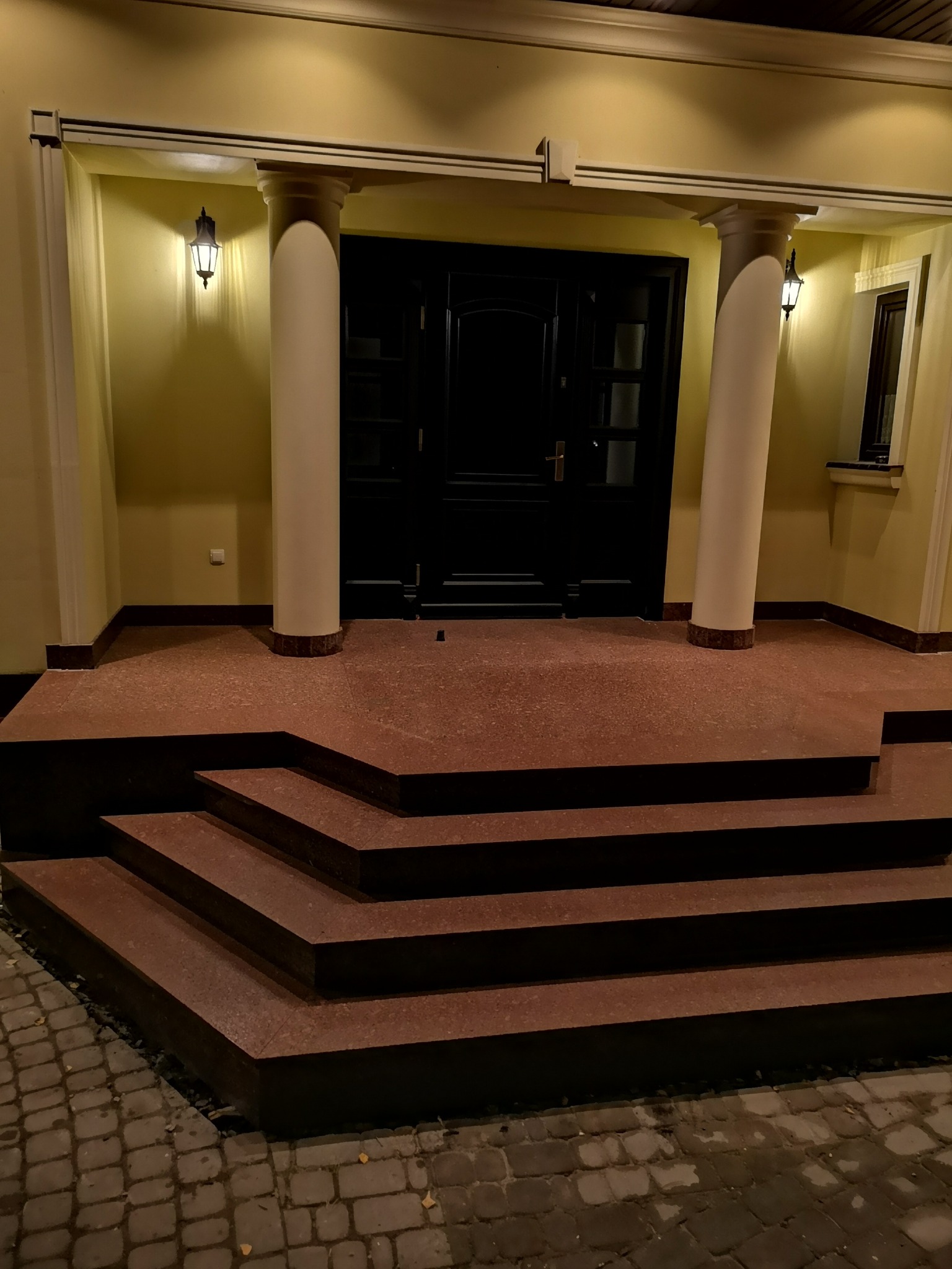 Wejście do domu z czterostopniowymi schodami w kolorze terakoty, podestem i dwoma kolumnami podtrzymującymi zadaszenie, oświetlone przez dwie kinkiety po bokach czarnych drzwi.