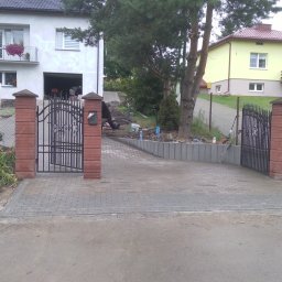 Układanie kostki brukowej Grzegorz Biernacki - Ułożona szara kostka brukowa na podjeździe do posesji z dwoma domami, widoczne otwarte bramy z ceglanymi słupkami, w tle prace ziemne i poziomica oparta o ogrodzenie.