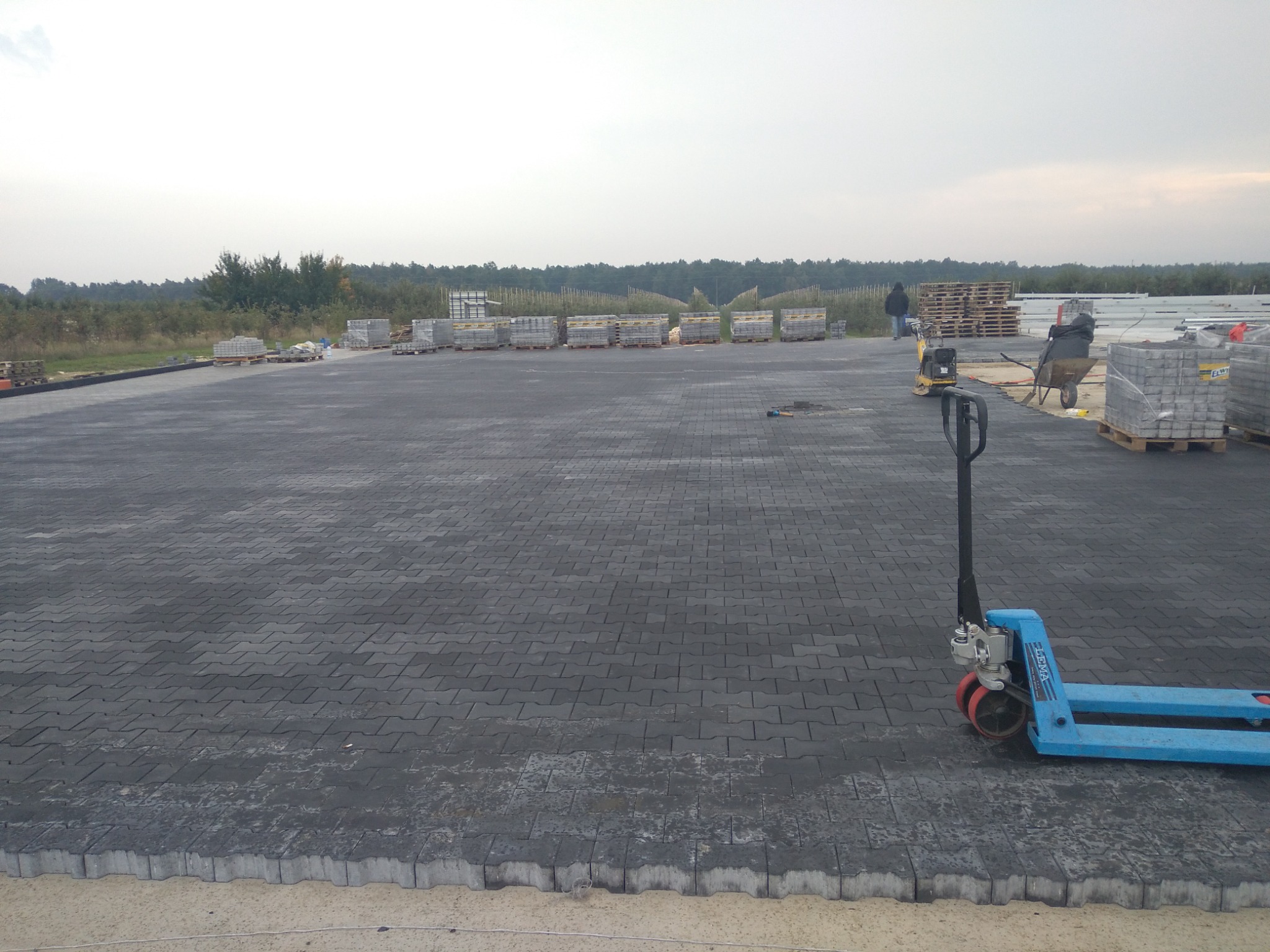 Rozległy plac z ułożoną szarą kostką brukową typu I, palety z kostką w tle, niebieski wózek paletowy z przodu po prawej stronie, częściowo widoczny obrzeże z kostki na piasku.