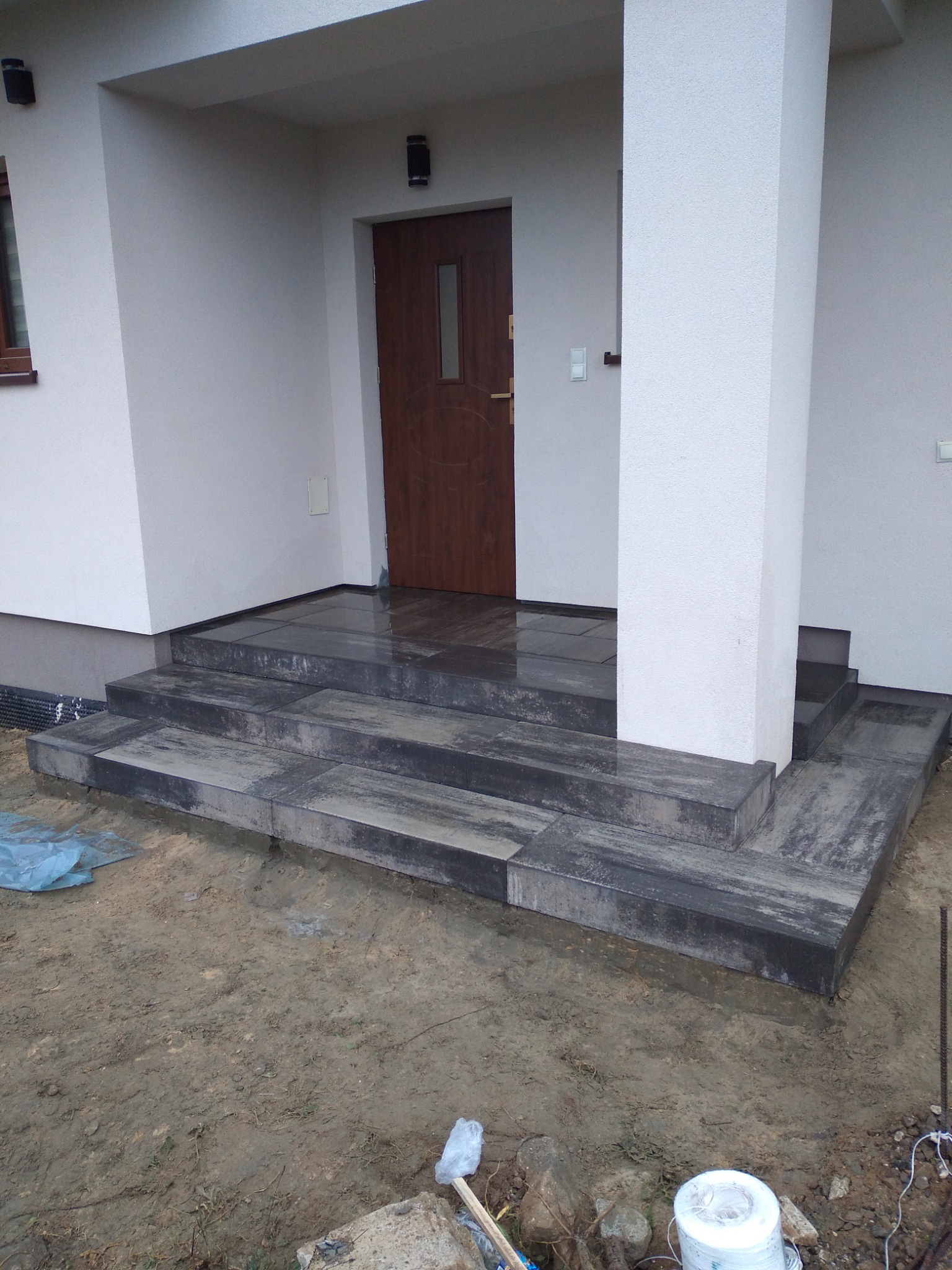 Schody wejściowe do domu wyłożone szarą kostką, widoczne świeże ślady po deszczu, teren wokół jeszcze niezagospodarowany, widoczne pozostałości po budowie.