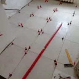 Układanie jasnych, kwadratowych płytek podłogowych w łazience z użyciem systemu poziomowania płytek (czerwone klipsy i kliny) oraz długiej poziomicy. Widoczne niedocięte płytki przy ścianach...