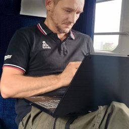 Sphaeris.pl Michał Grabarczyk - Mężczyzna w czarnej koszulce z logo Dakar, skupiony na pracy przy laptopie w pociągu PKP Intercity, z widokiem za oknem.