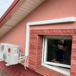 Serwis i montaż klimatyzacji Maciej Jezierski - Zewnętrzna jednostka klimatyzacji LG zamontowana na dachu skośnym domu jednorodzinnego, widoczne białe rury prowadzące do ściany, w oknie odbija się osoba.