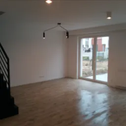 Wnętrze nowego, pustego pokoju z drewnopodobną podłogą, czarnymi schodami bez poręczy, białymi ścianami, grzejnikiem i nowoczesnym oświetleniem sufitowym.