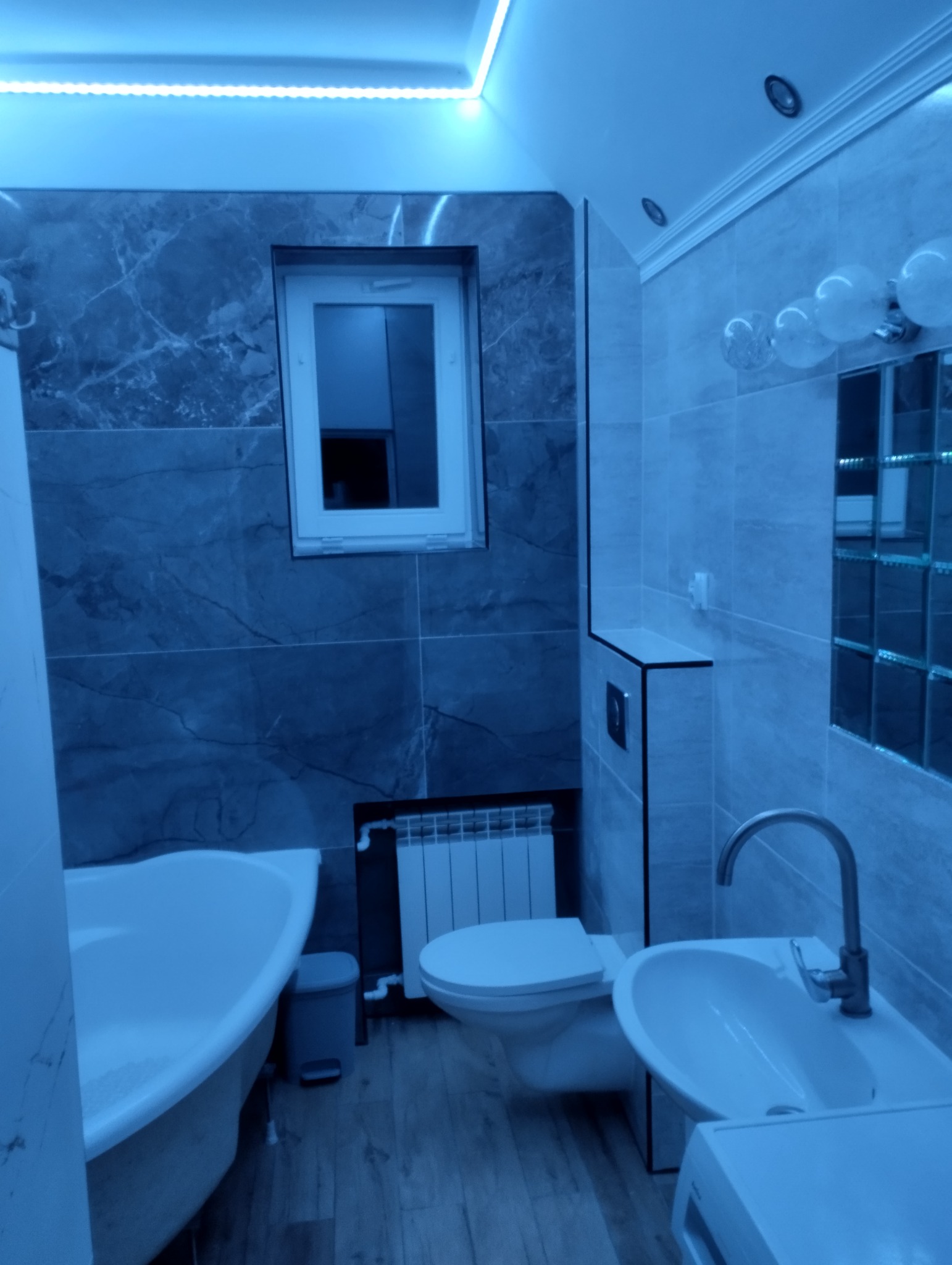 Nowoczesna łazienka w odcieniach szarości i błękitu z wanną, toaletą, umywalką i lustrem w stylu glamour, oświetlona listwą LED na suficie.