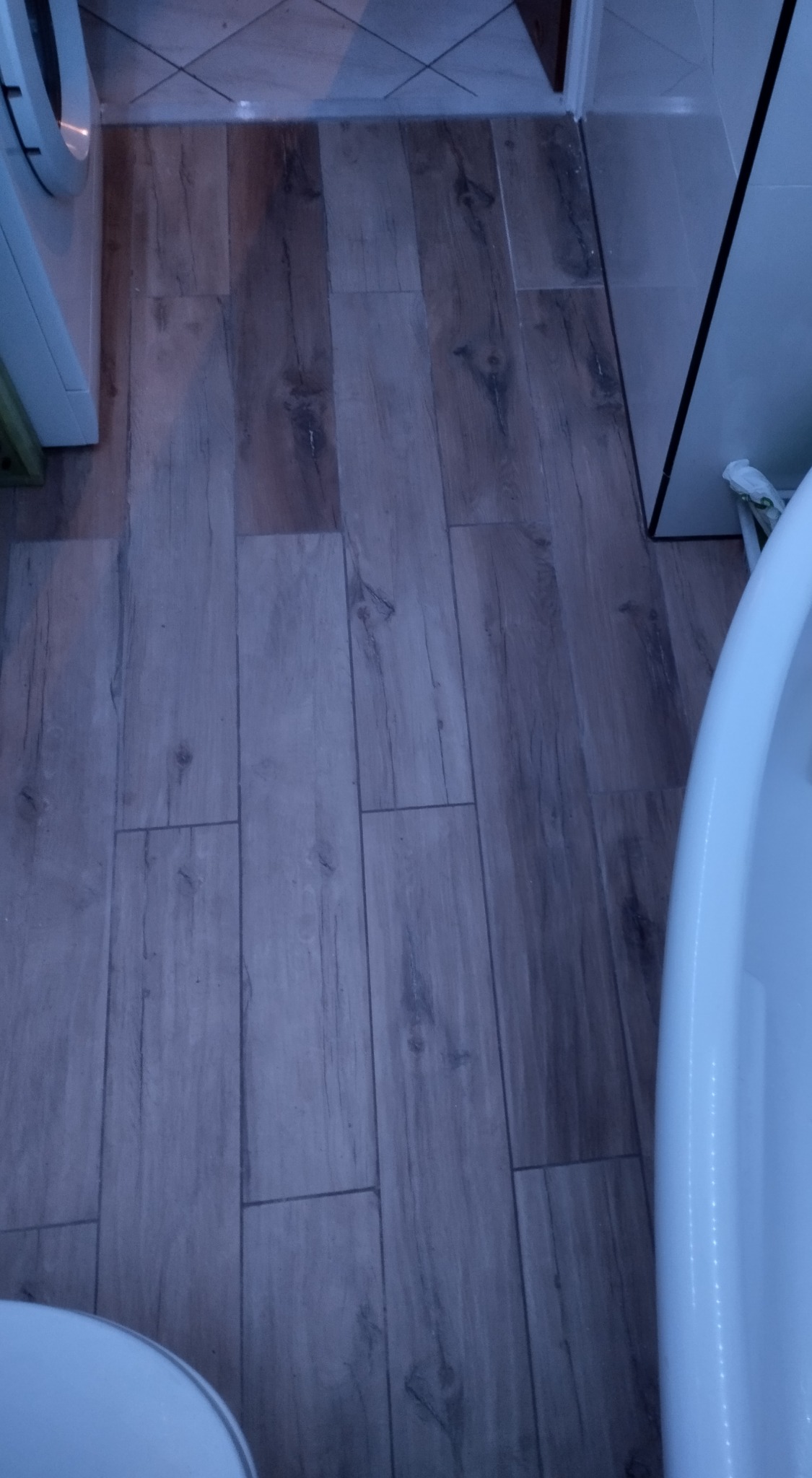 Ułożona podłoga z płytek imitujących drewno w łazience, widoczna pralka i fragment umywalki.