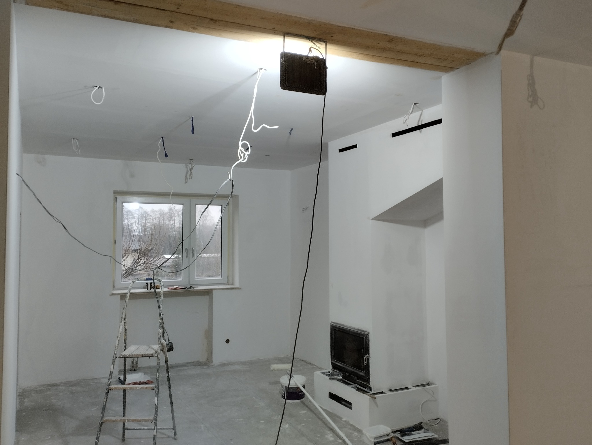 Wnętrze w trakcie prac przygotowawczych: białe ściany, surowy strop z drewnianych belek, widoczne przewody elektryczne, drabina malarska, piec kaflowy w trakcie obudowy.