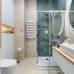 Nowoczesna łazienka z kabiną prysznicową ze szklanymi drzwiami, umywalką nablatową na drewnianym blacie, podświetlanym lustrem okrągłym, grzejnikiem drabinkowym i pralką w zabudowie.