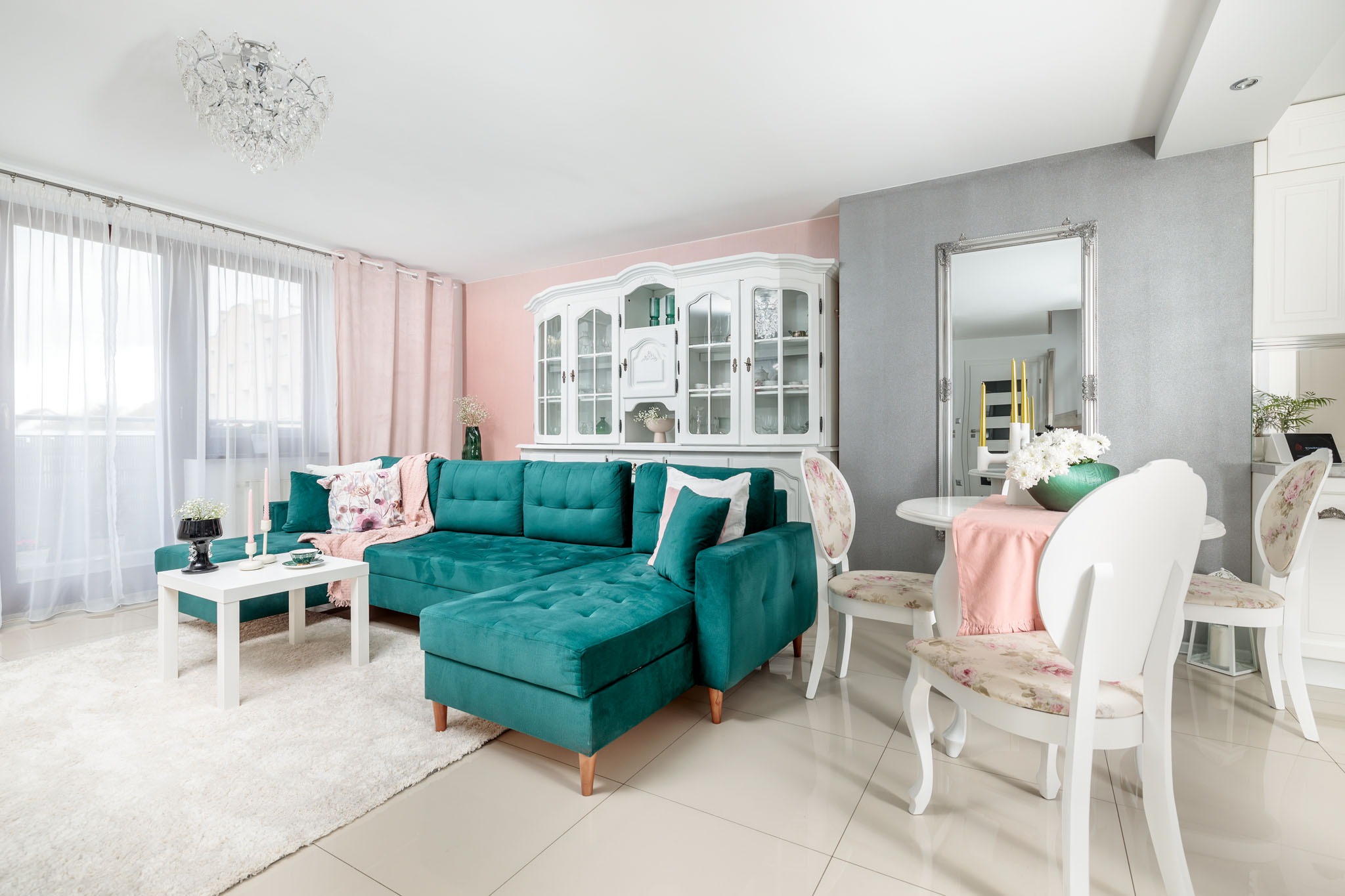 Turkusowy narożnik z aksamitną tapicerką, biały stolik kawowy, dywan shaggy i białe krzesła w pastelowym salonie z elementami różu i szarości.