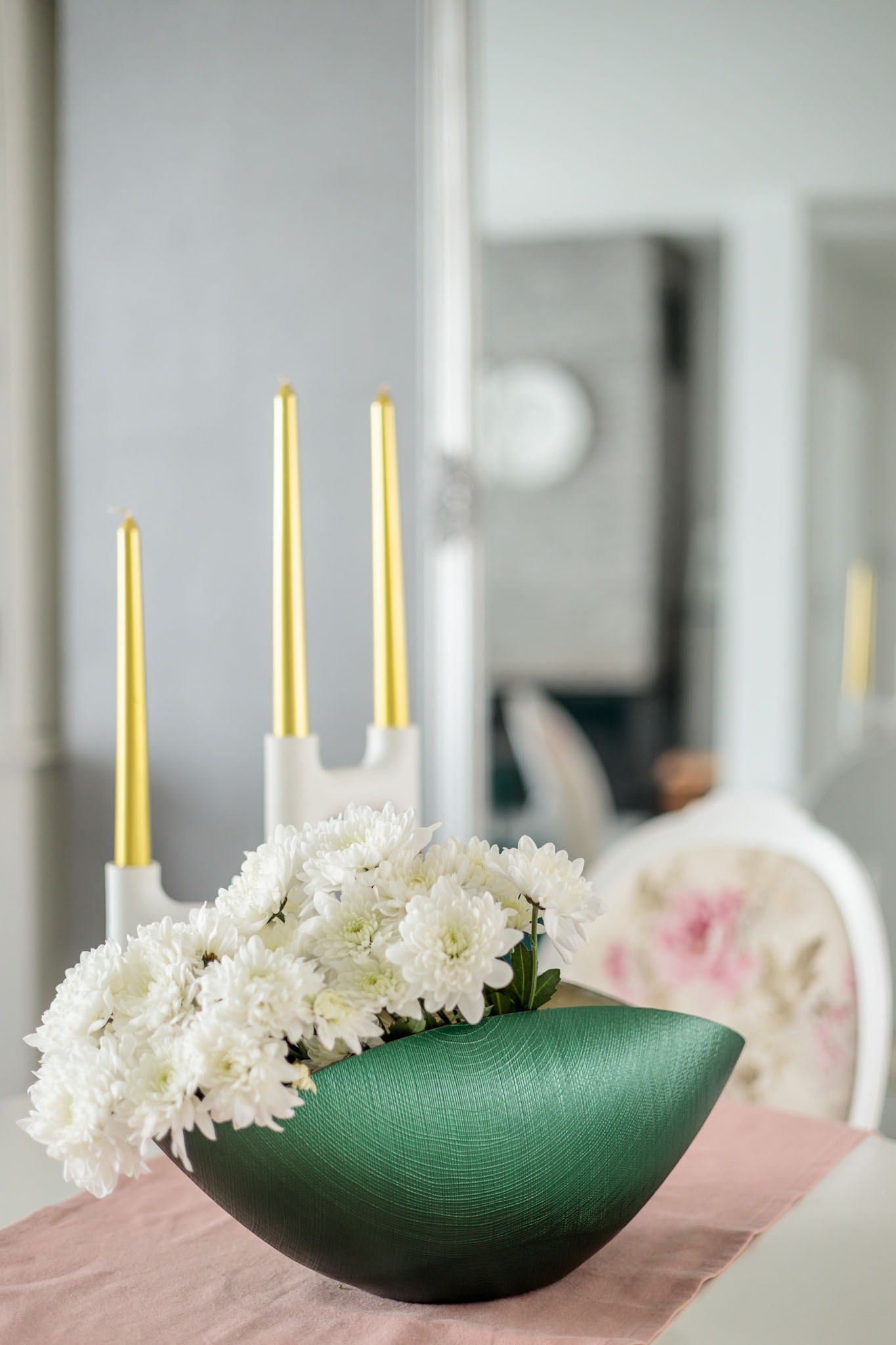 Dekoracja stołu: zielona misa z białymi chryzantemami i złotymi świecami na białym świeczniku, na różowym bieżniku, w tle krzesło z kwiatowym wzorem.