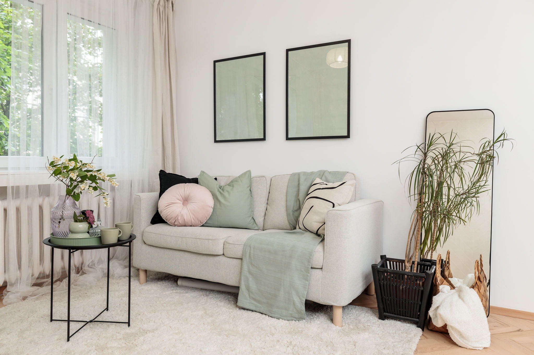 Jasny salon z szarą sofą, pastelowymi poduszkami, stolikiem z kwiatami i lustrem z rośliną, utrzymany w neutralnej kolorystyce.