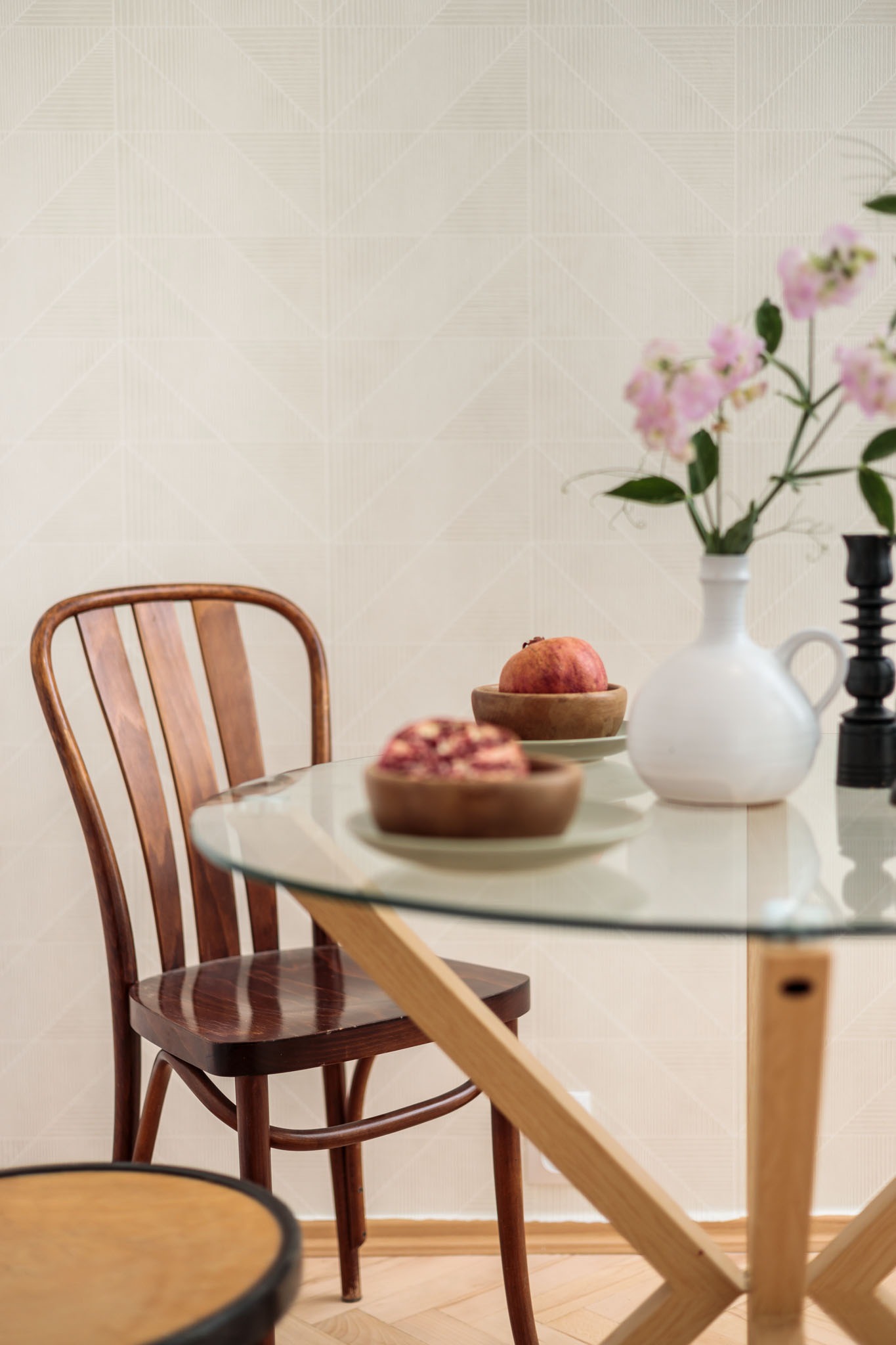 Szczecińskie wnętrze z okrągłym szklanym stołem, drewnianym krzesłem giętym, owocami w drewnianych miseczkach i białym wazonem z różowymi kwiatami na tle ściany w geometryczne wzory.