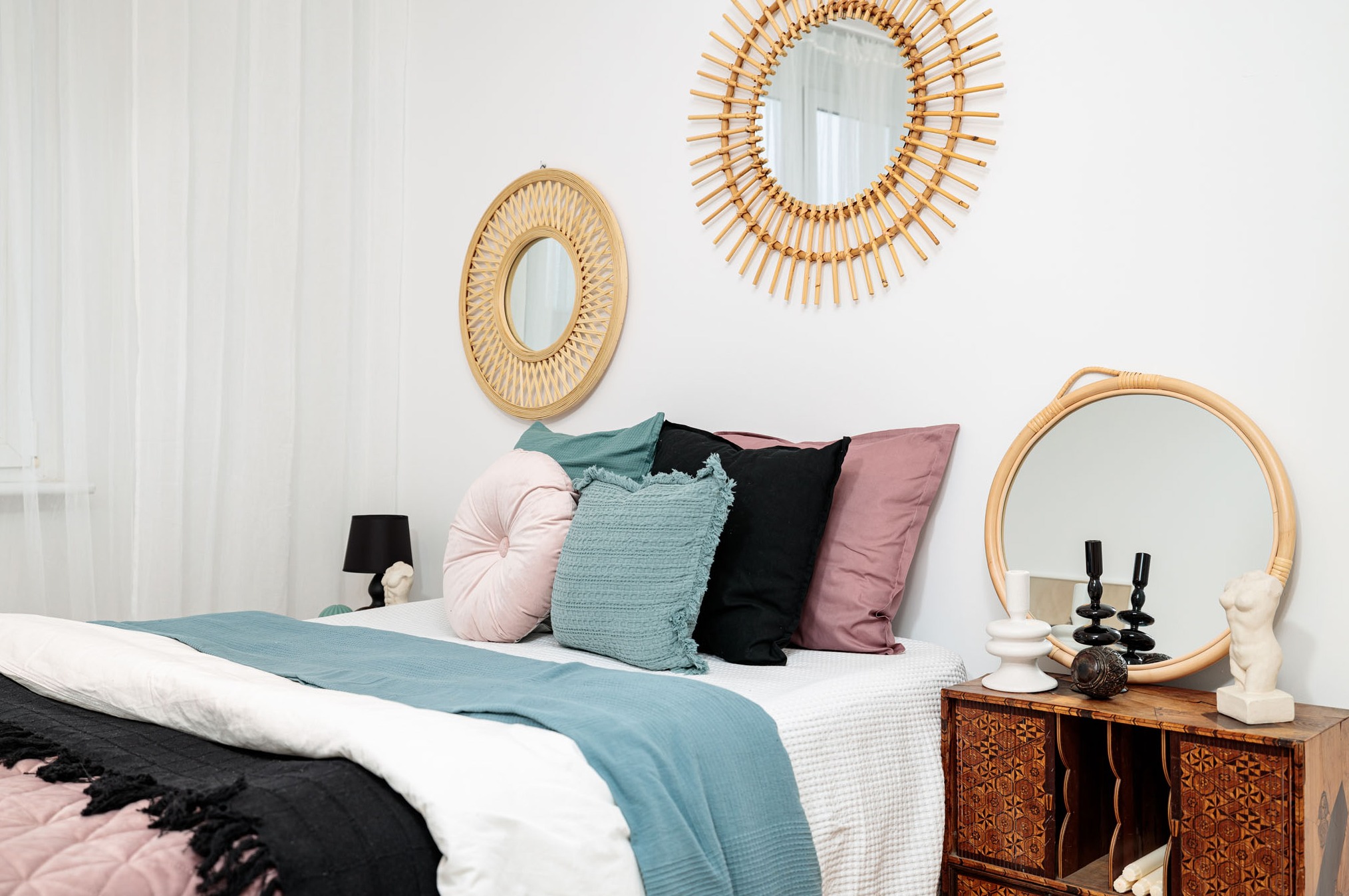 Jasna sypialnia z łóżkiem, dekoracyjnymi poduszkami w odcieniach różu, czerni i błękitu, trzema okrągłymi lustrami w rattanowych ramach oraz drewnianą komodą z rzeźbieniami.