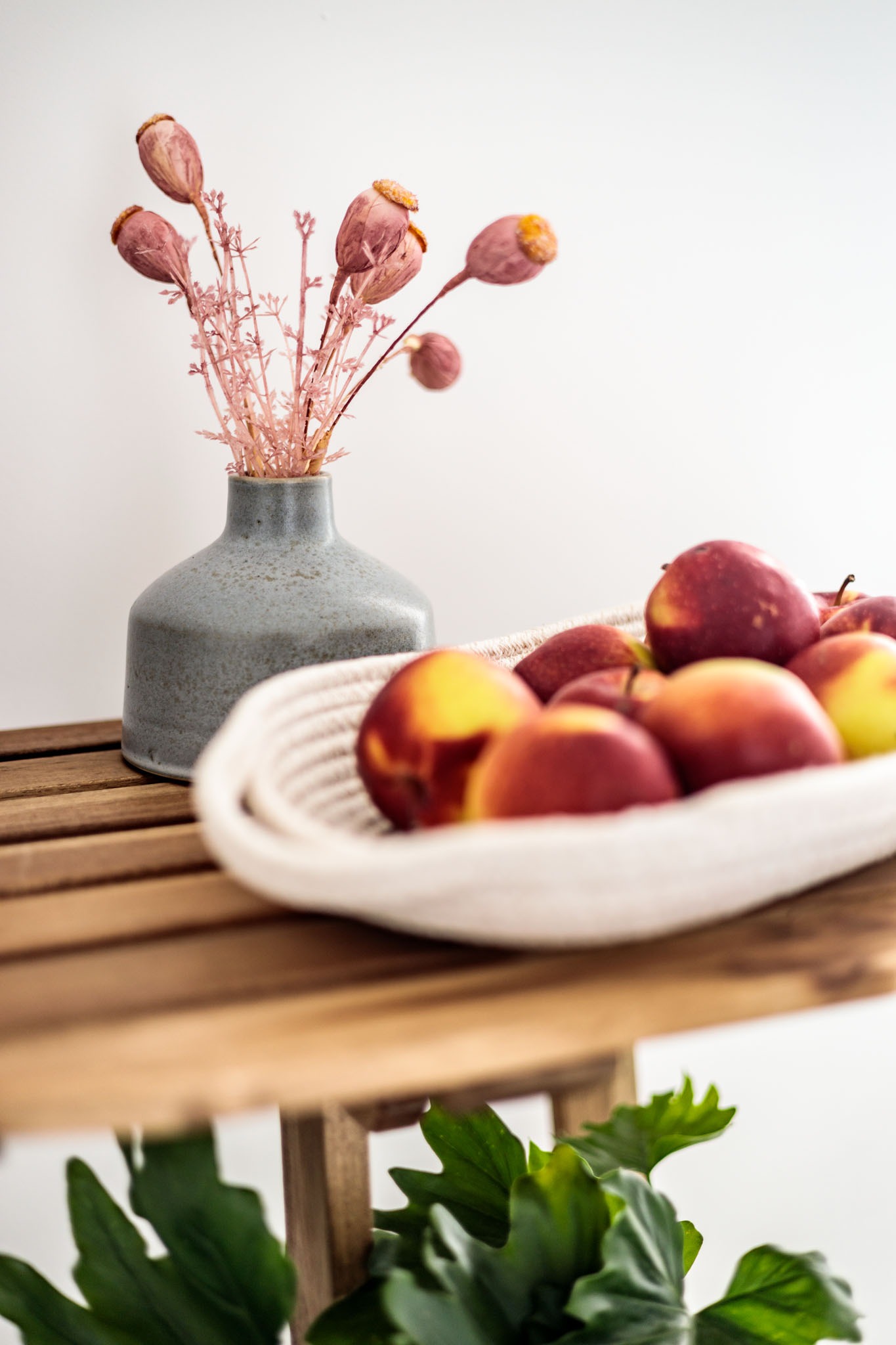 Martwa natura z brzoskwiniami w plecionym koszyku, szary wazon z suszonymi makówkami i zieloną rośliną na drewnianym stoliku.