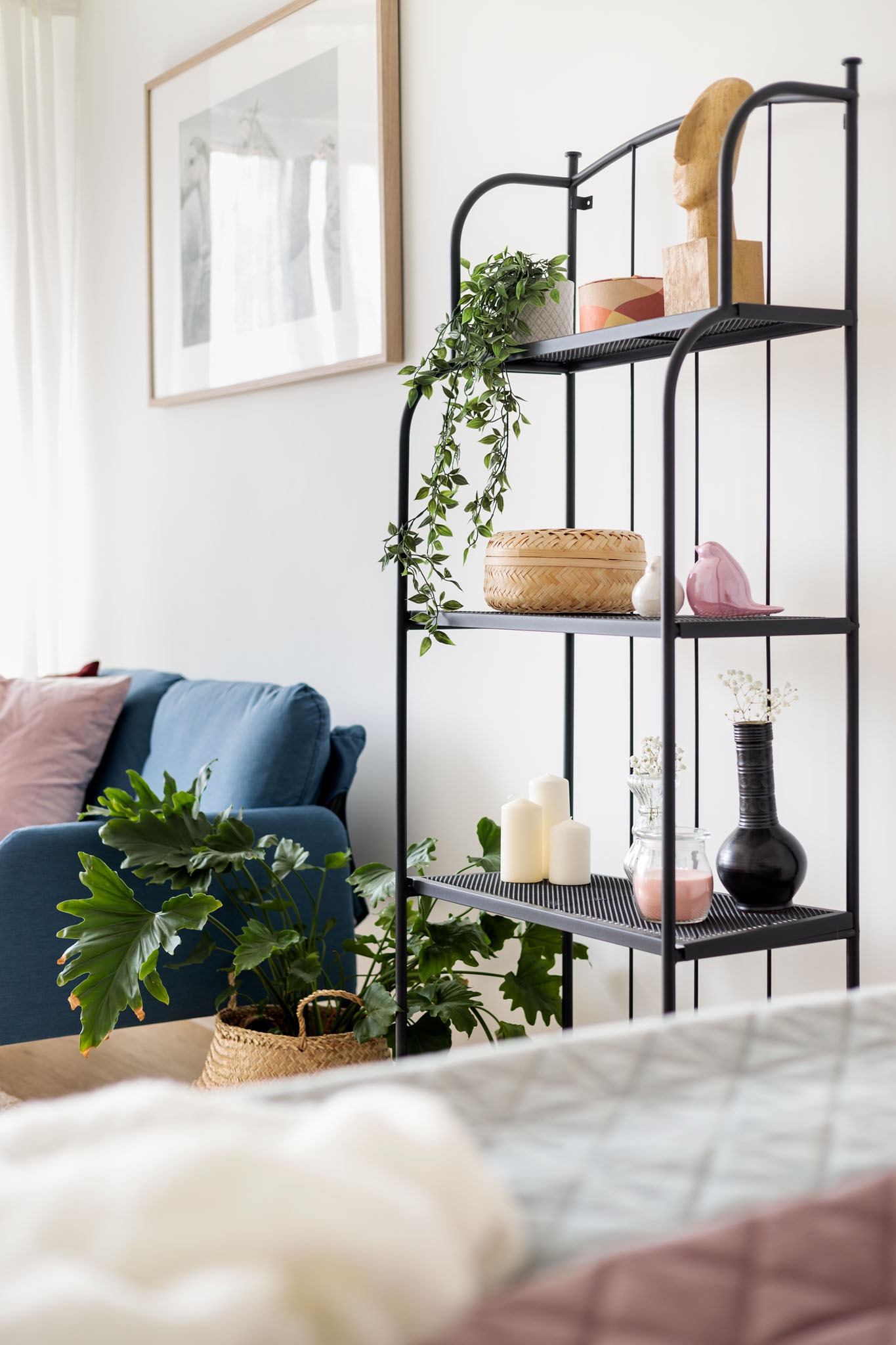 Czarny, metalowy regał z dekoracjami: rośliny doniczkowe, świece, wazoniki, ceramiczna figurka ptaka, drewniana rzeźba, na tle błękitnej sofy i grafiki w ramie.