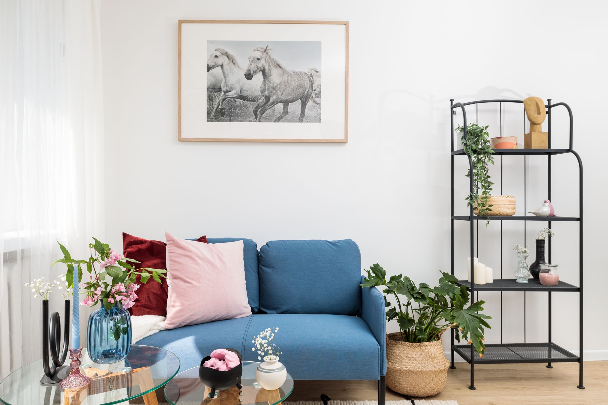 Jasne wnętrze pokoju dziennego z błękitną sofą, szklanym stolikiem kawowym, metalowym regałem z dekoracjami i obrazem koni na ścianie.