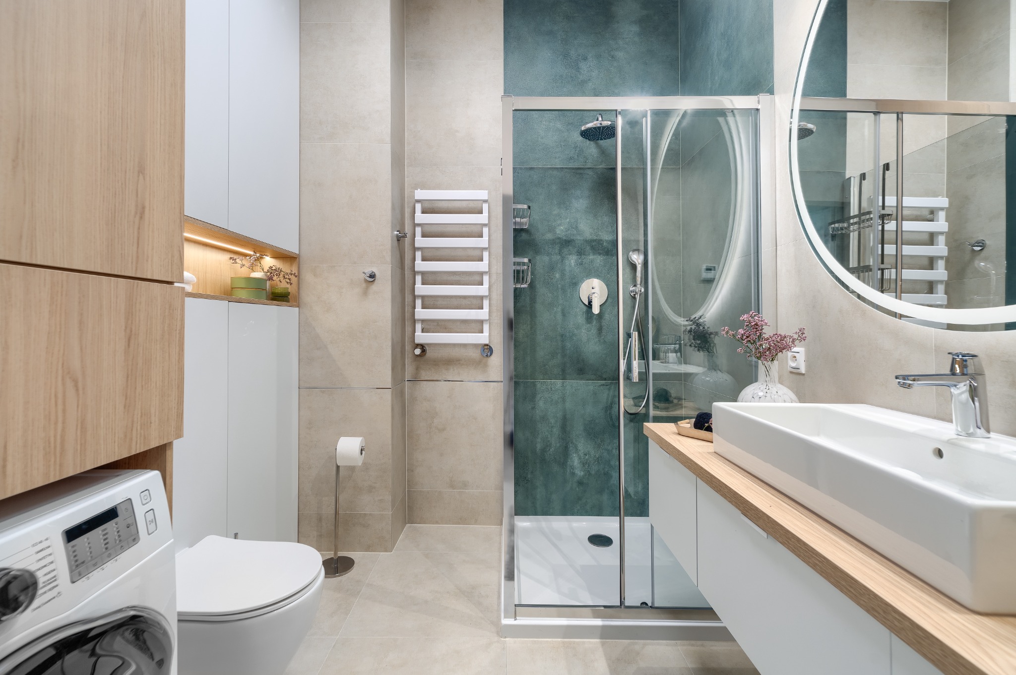 Nowoczesna łazienka z kabiną prysznicową ze szklanymi drzwiami, umywalką nablatową na drewnianym blacie, podświetlanym lustrem okrągłym, grzejnikiem drabinkowym i pralką w zabudowie.