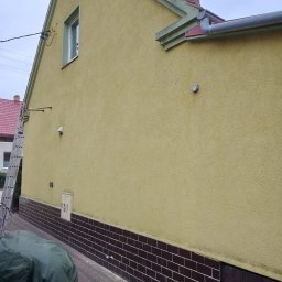 Malowanie dachów Bartlomiej Świątek - Żółta elewacja domu z brązową podmurówką. Aluminiowa drabina oparta o ścianę. Widoczny fragment rynny i okna. Częściowo widoczna postać w kurtce.