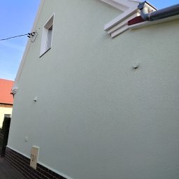 Malowanie dachów Bartlomiej Świątek - Jasnozielona, odnowiona elewacja domu jednorodzinnego z brązową podmurówką i białą stolarką okienną. Widoczny fragment dachu i rynny.