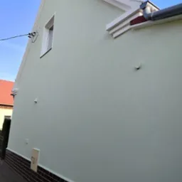 Jasnozielona, odnowiona elewacja domu jednorodzinnego z brązową podmurówką i białą stolarką okienną. Widoczny fragment dachu i rynny.