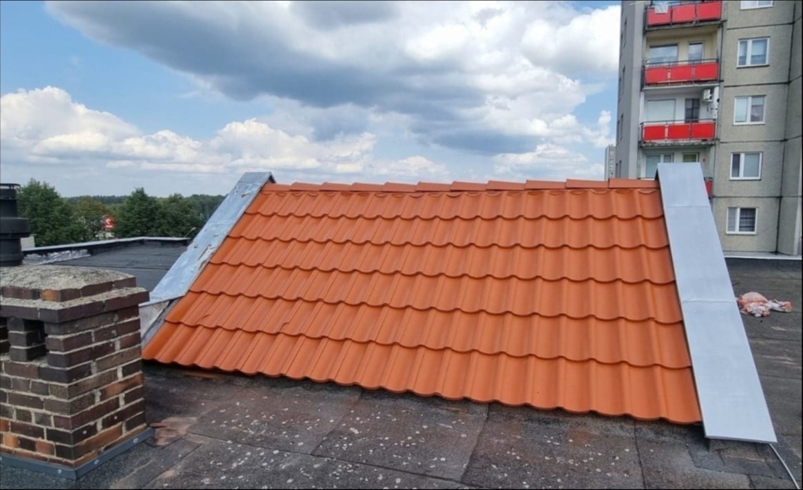 Czerwona dachówka na dachu budynku wielorodzinnego, widoczny ceglany komin i metalowe obróbki blacharskie.