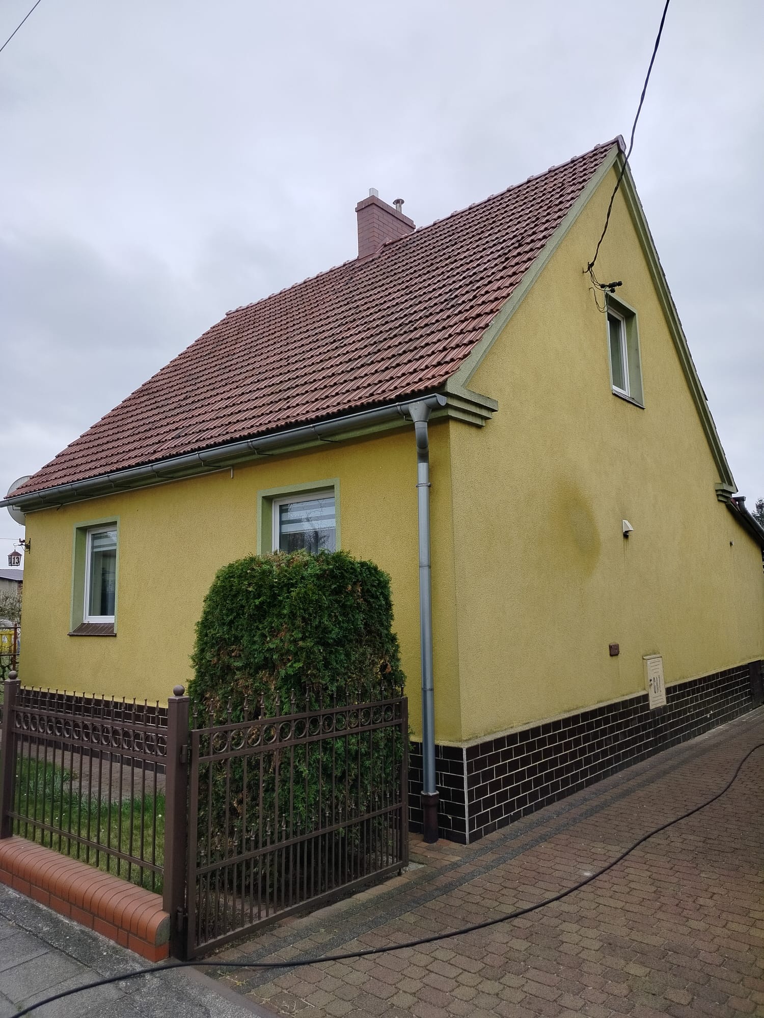 Żółty dom z brązowym dachem i ciemną podmurówką, widok z boku, z widoczną rynną i fragmentem ogrodzenia. Pochmurne niebo w tle.