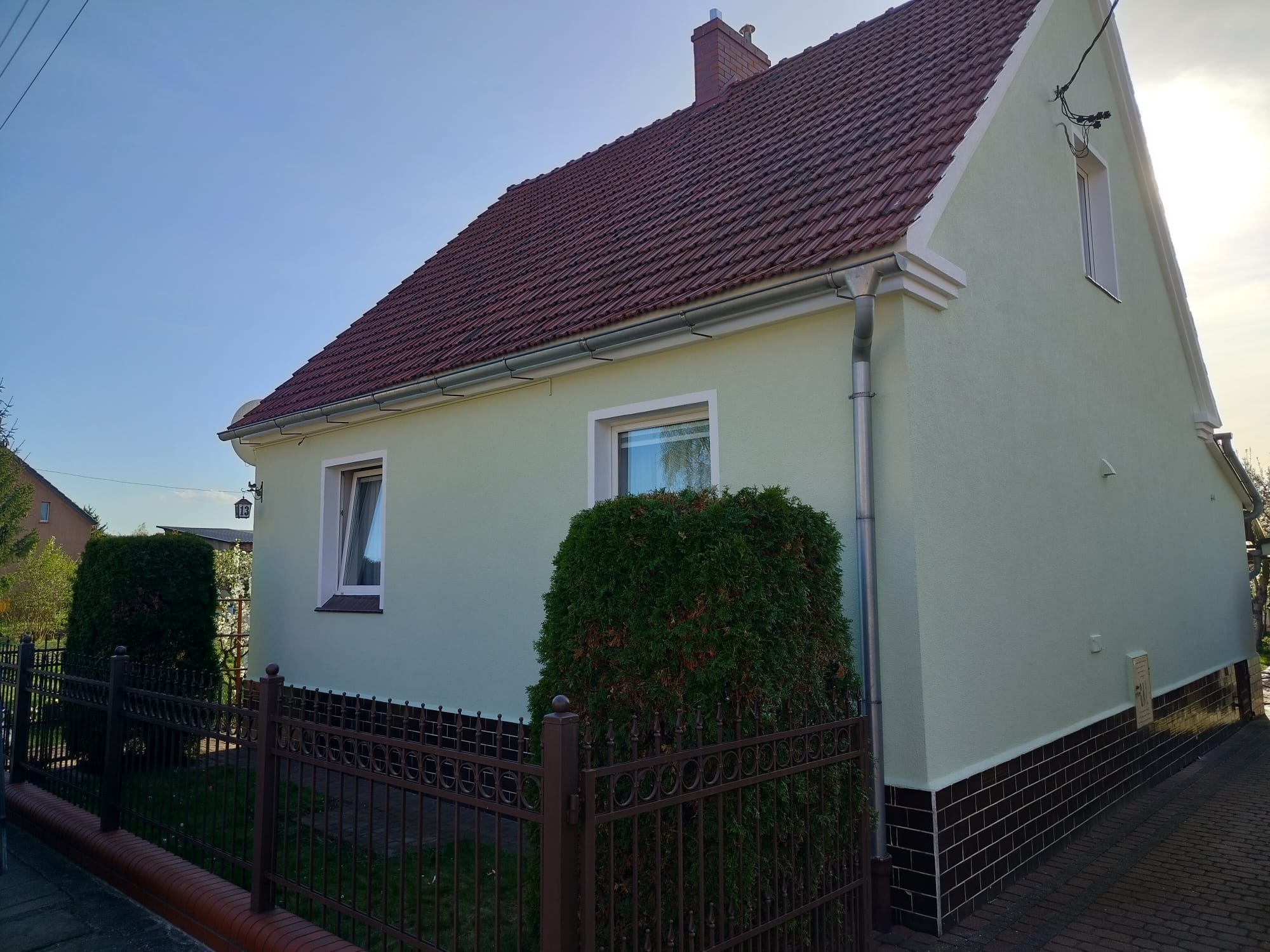 Odnowiona elewacja domu z brązowym dachem i rynnami, widok z boku, ogrodzenie z metalowymi przęsłami i zielone krzewy w ogrodzie.