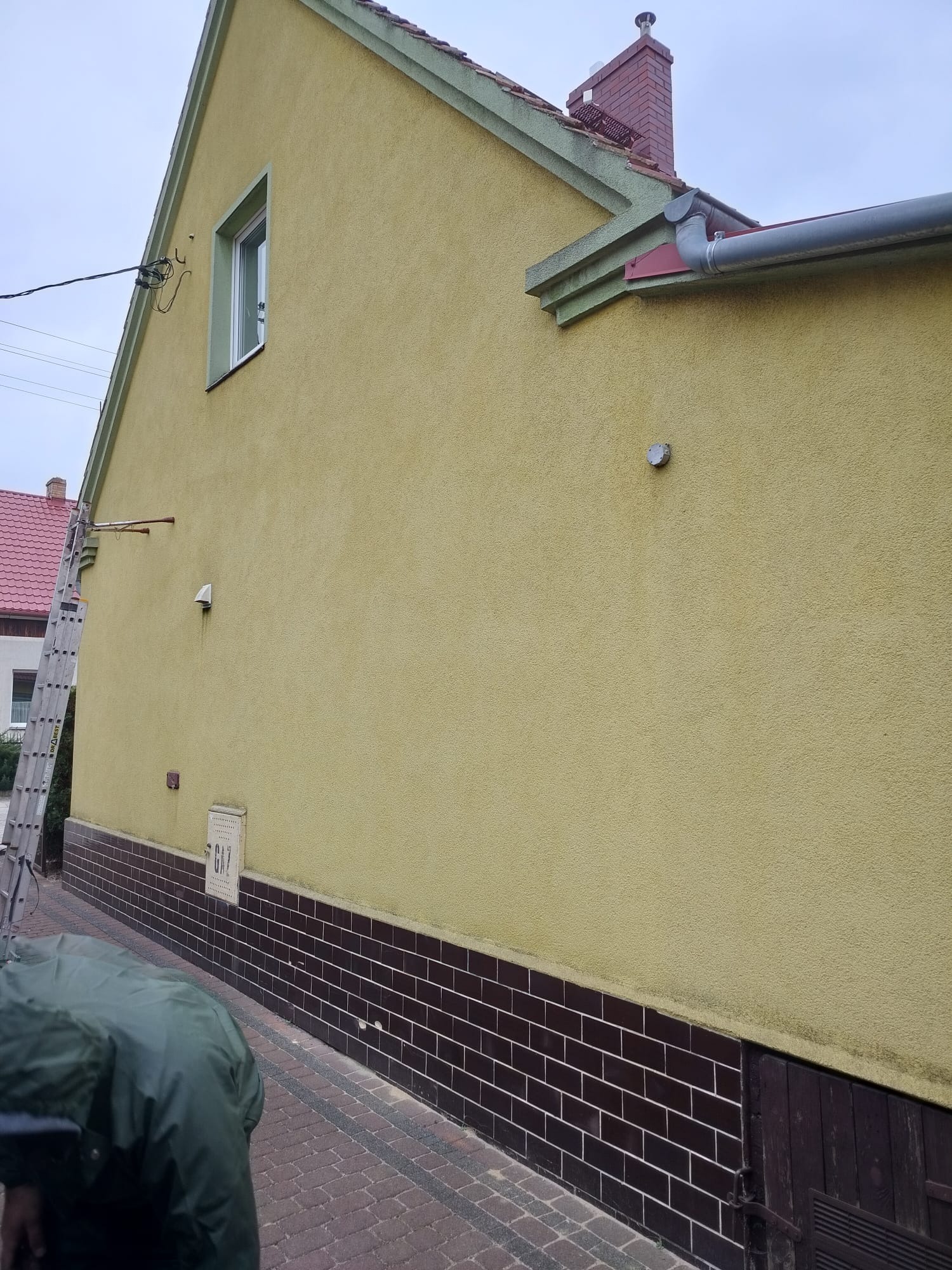 Żółta elewacja domu z brązową podmurówką. Aluminiowa drabina oparta o ścianę. Widoczny fragment rynny i okna. Częściowo widoczna postać w kurtce.