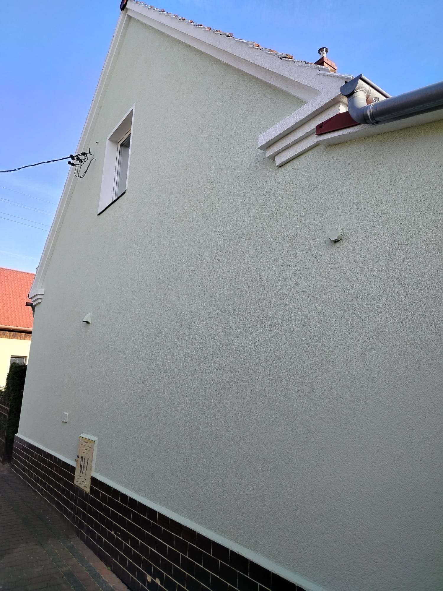 Jasnozielona, odnowiona elewacja domu jednorodzinnego z brązową podmurówką i białą stolarką okienną. Widoczny fragment dachu i rynny.