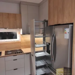 Nowoczesna kuchnia z drewnianymi i szarymi szafkami, wysuwana szafka cargo obok lodówki Samsung, płyta grzewcza na blacie.