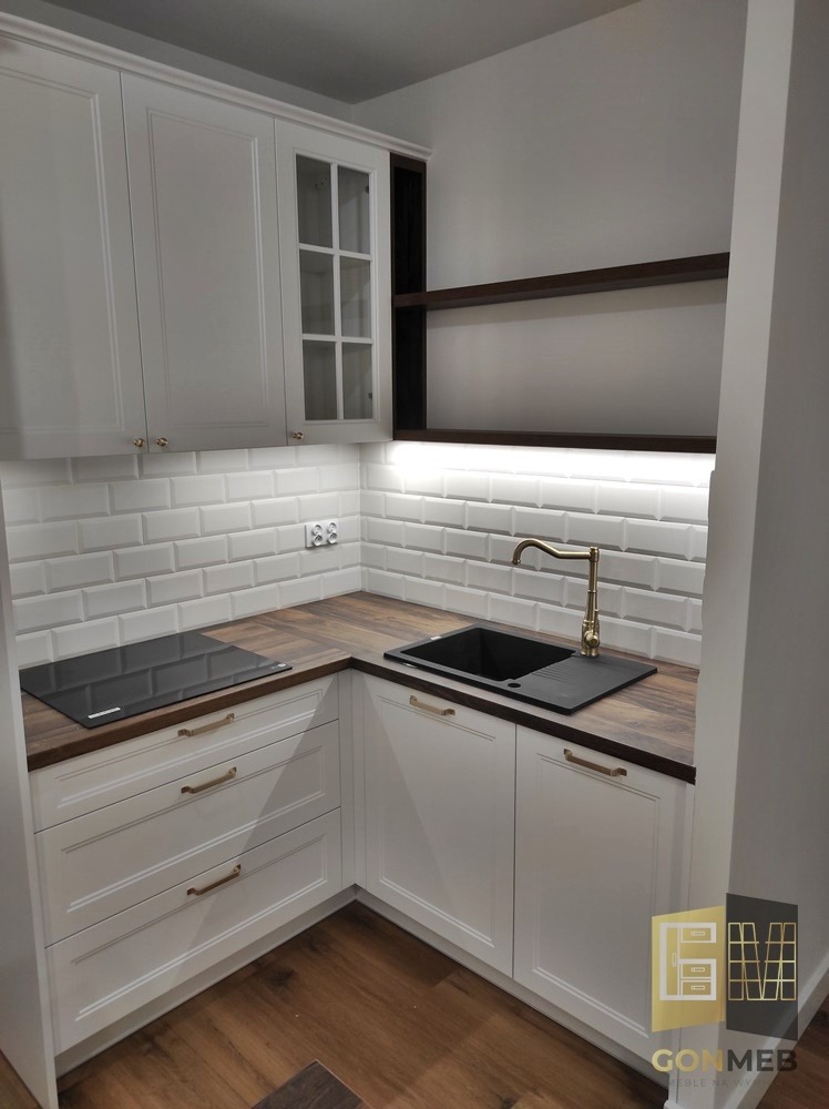 Biała, narożna zabudowa kuchenna z drewnianym blatem, złotą armaturą, płytkami typu metro i otwartymi półkami, oświetlona listwą LED podszafkową, z widocznym logo firmy w prawym dolnym rogu.