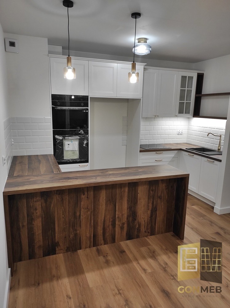 Nowoczesna kuchnia z białymi szafkami, drewnianym blatem i wyspą w odcieniach brązu. Nad wyspą wiszą dwie lampy o różnym designie. Na ścianie białe płytki imitujące cegłę.