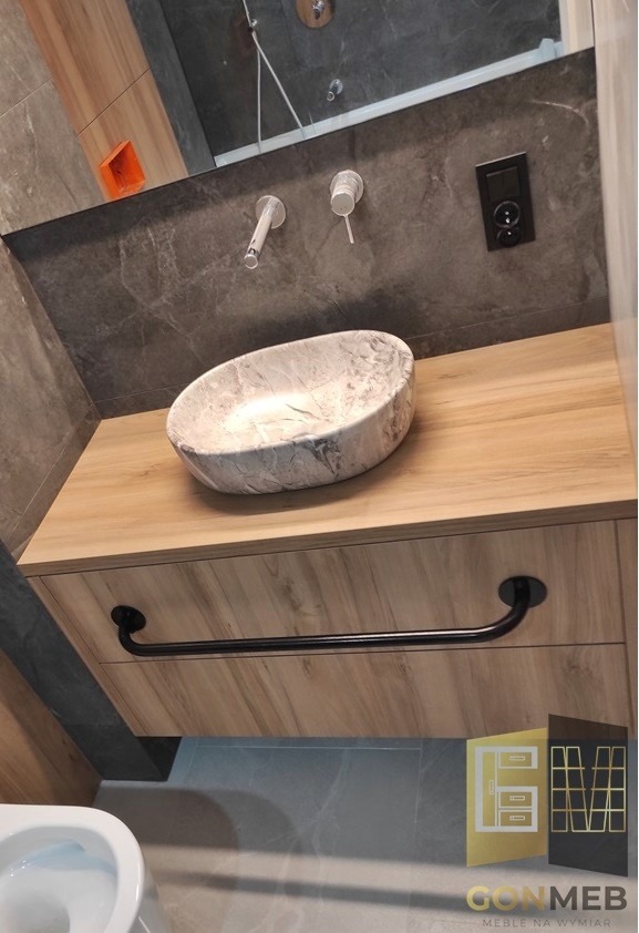 Łazienka z marmurową umywalką nablatową o owalnym kształcie, zamontowaną na szafce z drewnopodobnym blatem i czarnym, minimalistycznym uchwytem. Na ścianie widoczna armatura i czarny panel...