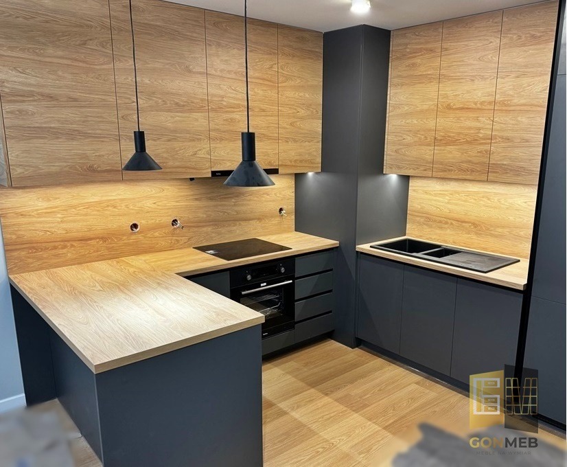 Nowoczesna kuchnia z drewnianymi frontami szafek górnych i blatem, kontrastująca z grafitowymi szafkami dolnymi i ścianą, oświetlona wiszącymi lampami w kształcie stożków.