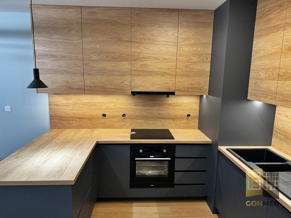 Nowoczesna kuchnia z drewnianymi frontami szafek górnych i blatem, połączona z grafitowymi szafkami dolnymi i czarnym okapem, oświetlona punktowo pod szafkami.