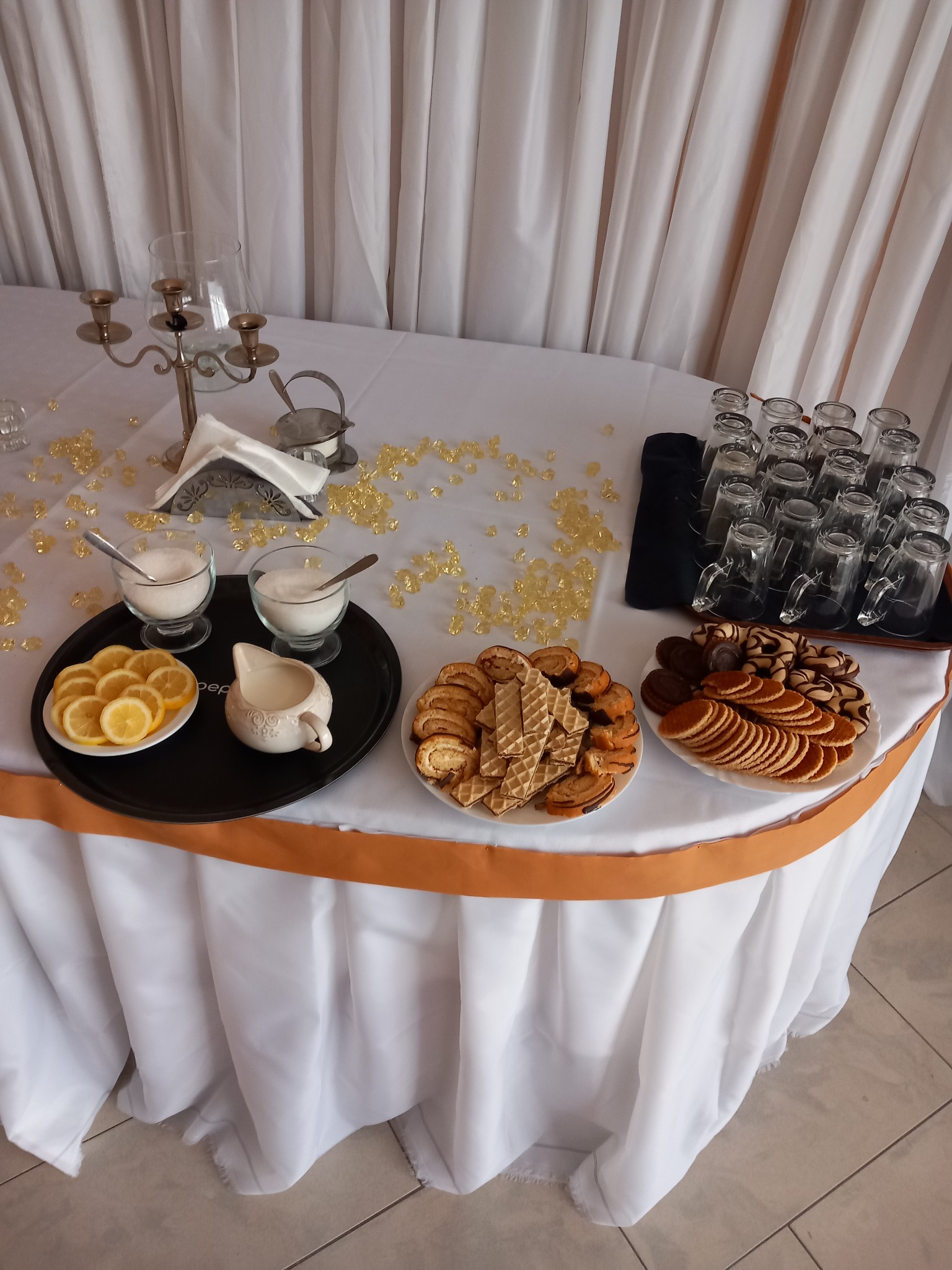 Stół z białym obrusem udekorowany złotymi elementami, na którym znajdują się talerze z ciastkami, wafle, cytryny, dzbanuszek na mleko, cukiernice ze srebrnymi łyżeczkami oraz szklanki na tacy.
