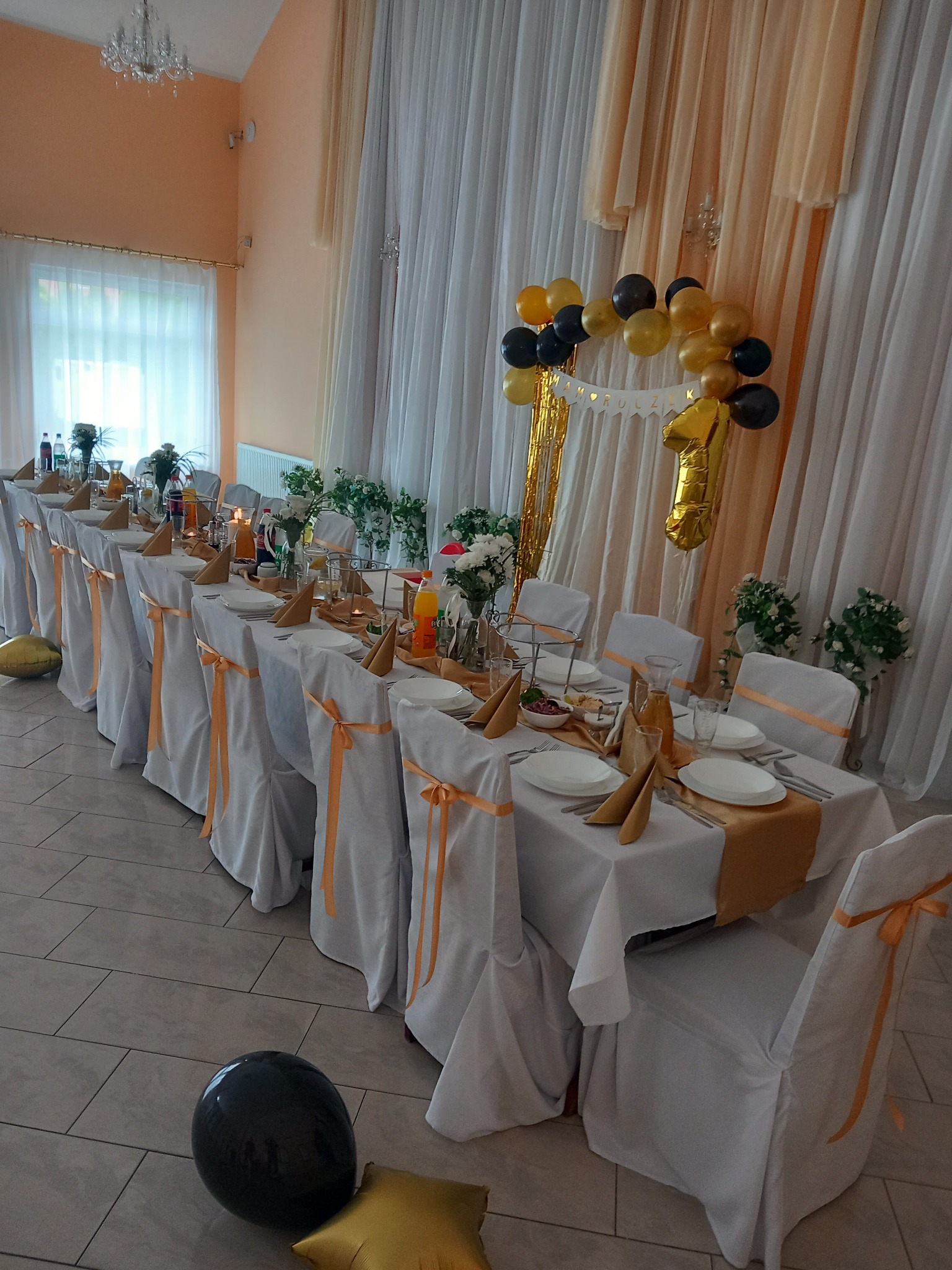 Uroczysta dekoracja stołu w Maksymilianowie na przyjęcie urodzinowe z białymi obrusami, złotymi akcentami, balonami i kwiatami.