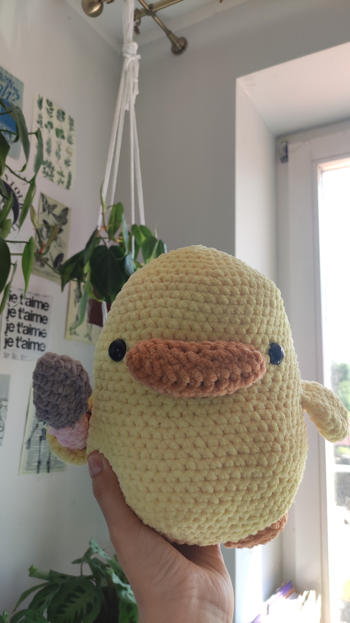 Ręcznie robiona, żółta, pluszowa kaczka z brązowym dziobem i oczami z guzików, trzymana w dłoni, w tle okno i rośliny doniczkowe