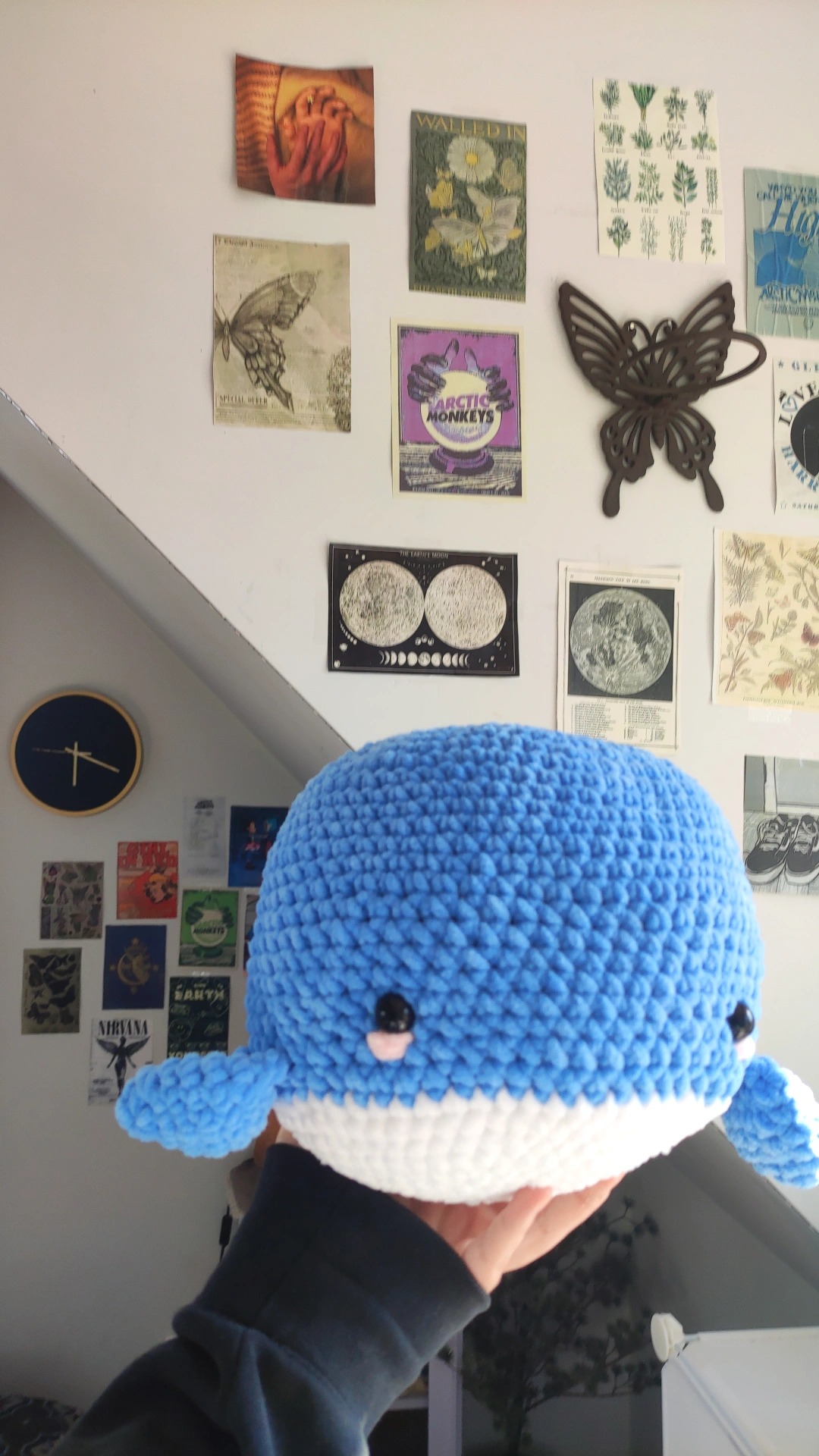 Ręcznie robiona, niebiesko-biała, pluszowa maskotka wieloryba, trzymana w dłoni, z widocznym tłem ściany udekorowanej plakatami i zegarem.